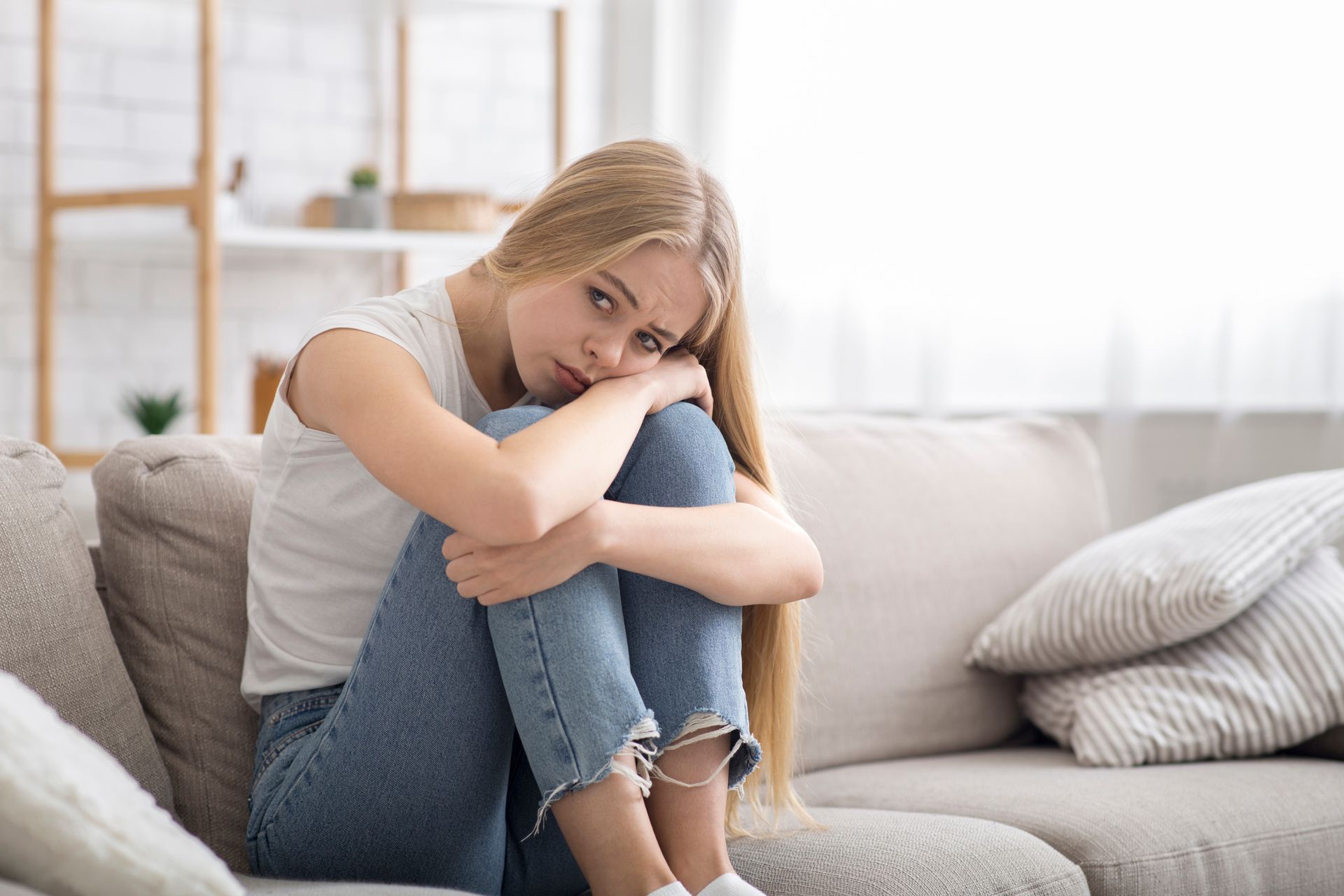 Woman sitting curled up on a couch, resting her head on her knees in a bright living room