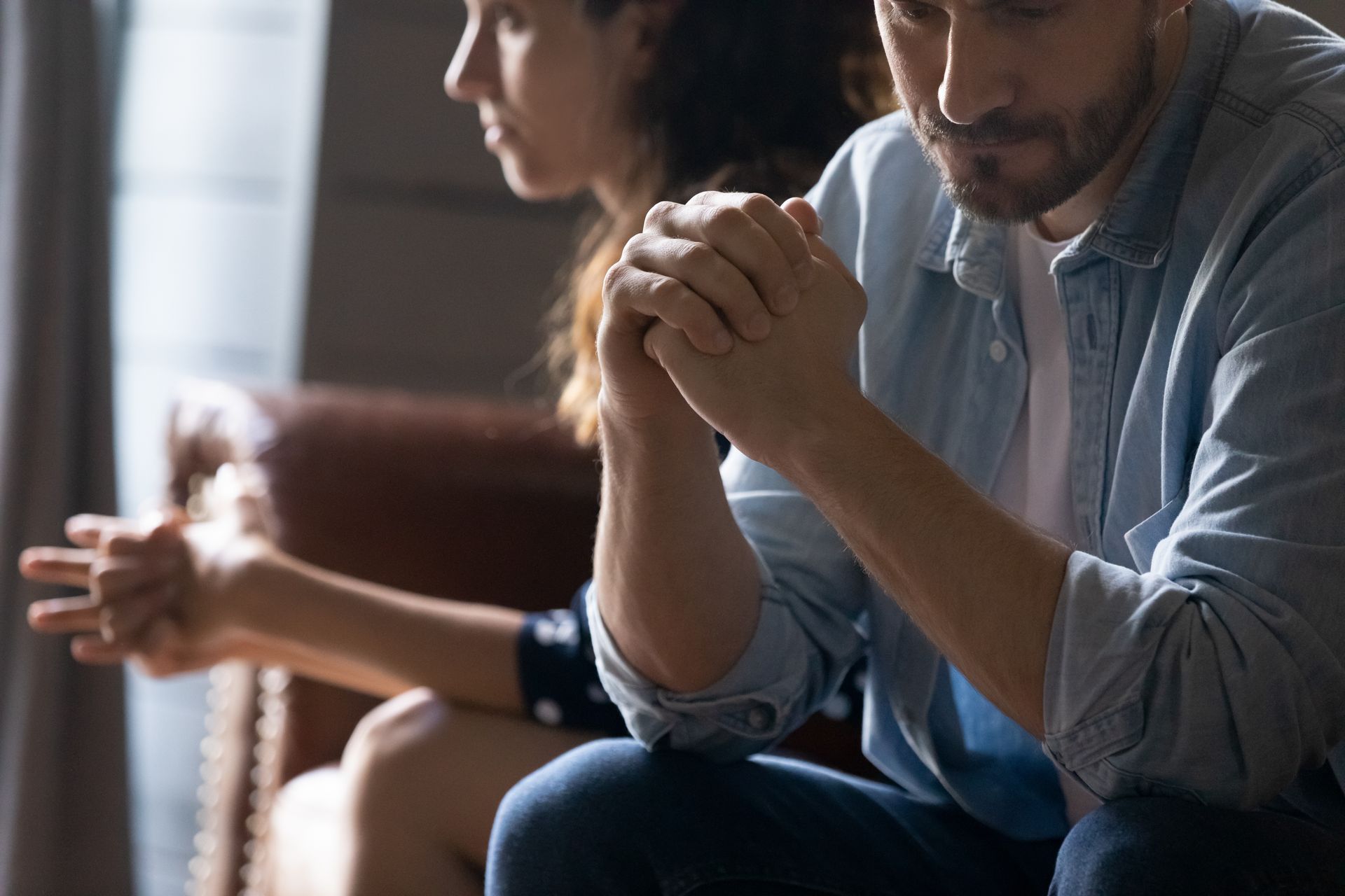 Two people sit with clasped hands in a softly lit room, one looking down in thought.
