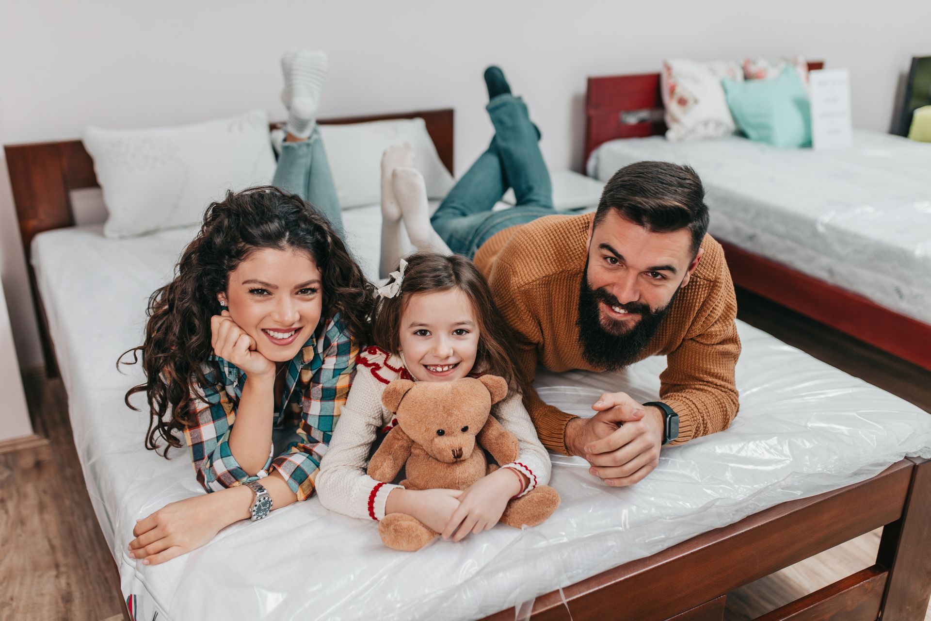 Three people lying on a bed, smiling with a teddy bear in a bright bedroom