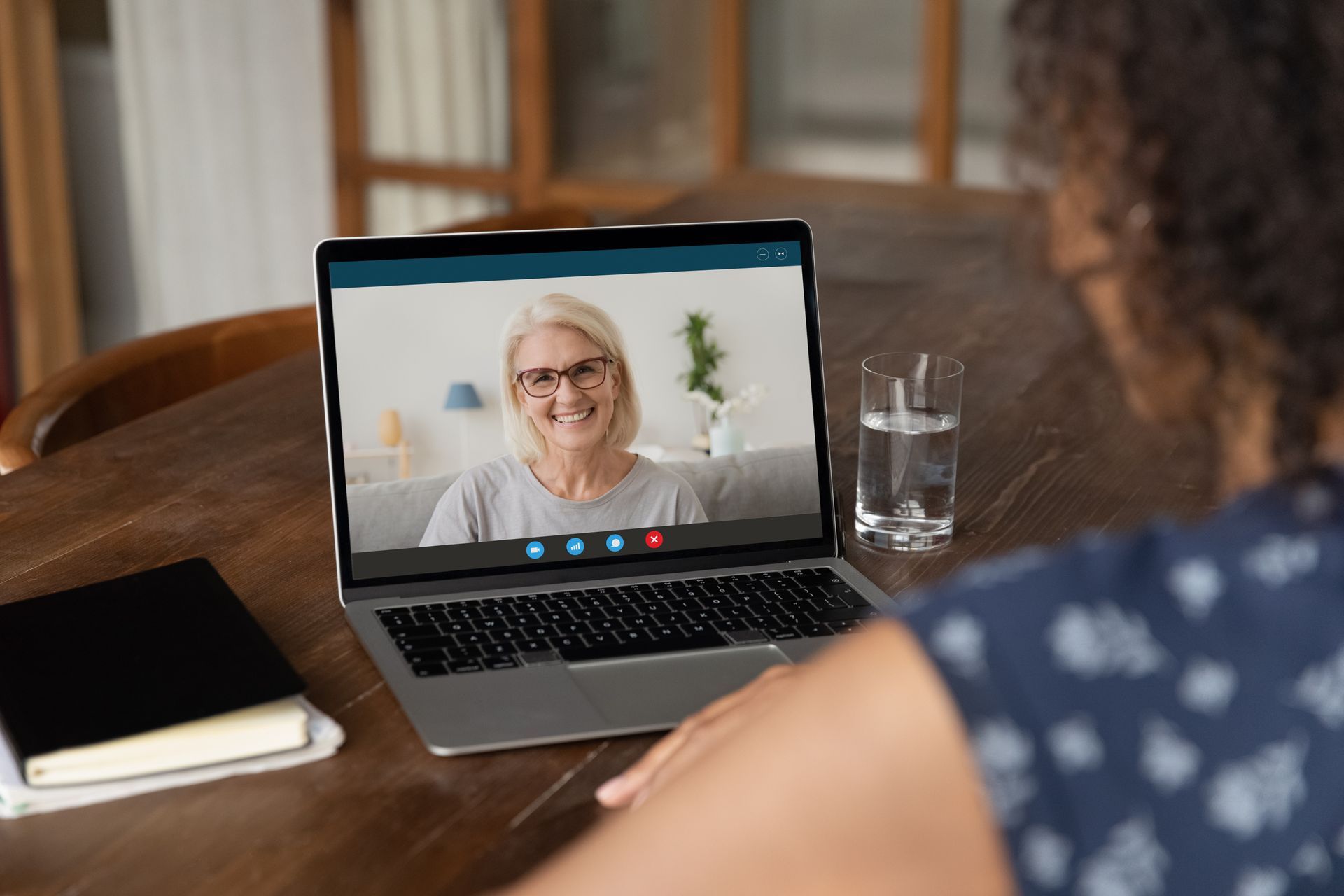 Video call on a laptop with a smiling woman, viewed from behind at a wooden table.