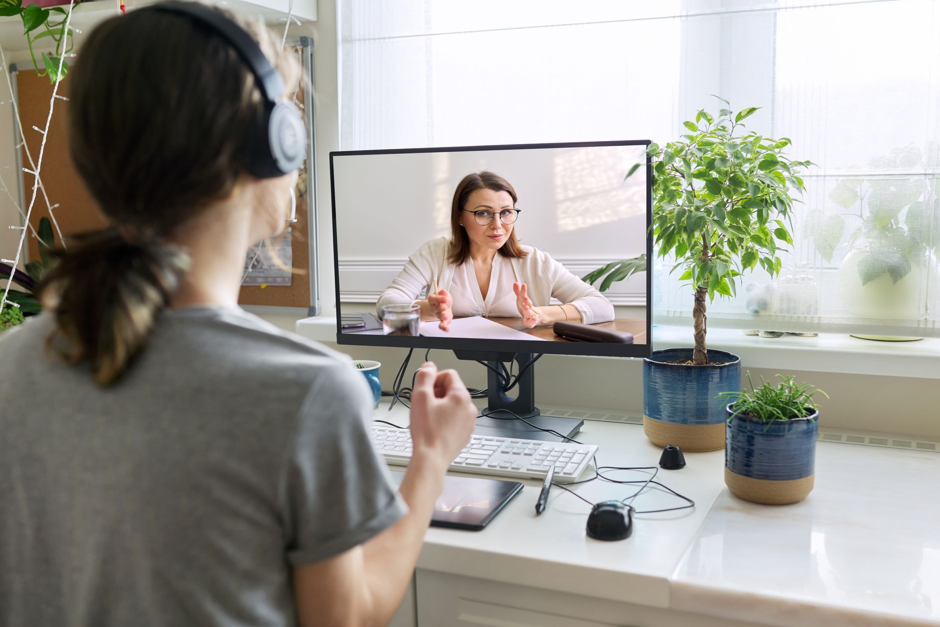 Person on headset video calling a woman at a desk, with plants and a bright office window.