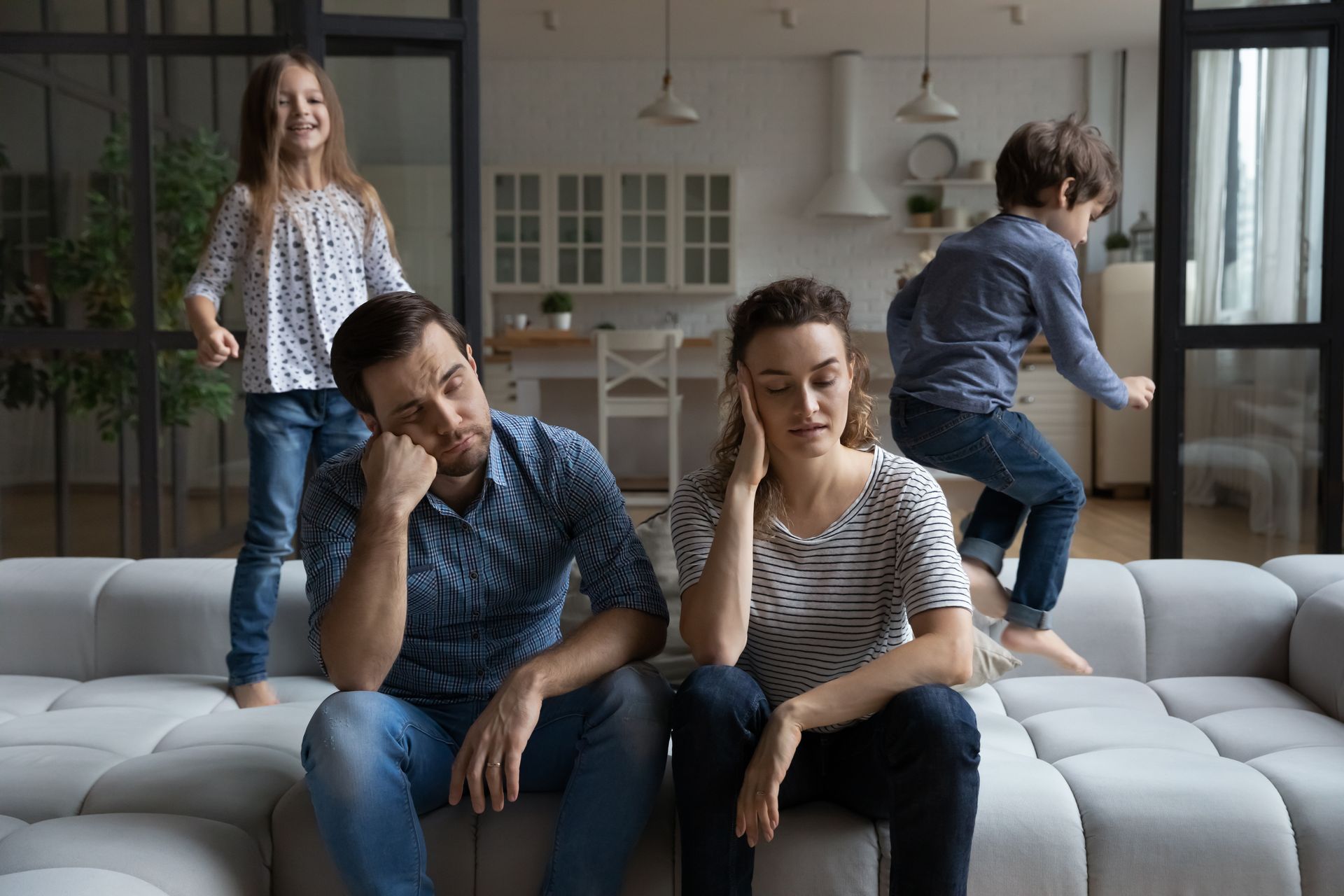 Two adults sit on a couch looking tired while two children play behind them in a living room