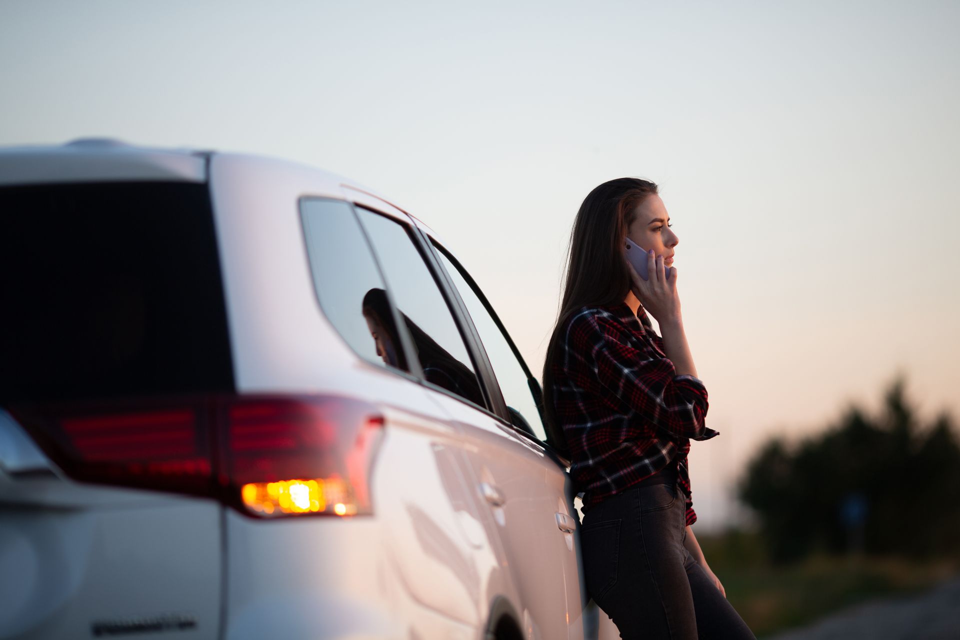 Woman standing beside a white SUV at sunset, talking on a phone on a roadside.