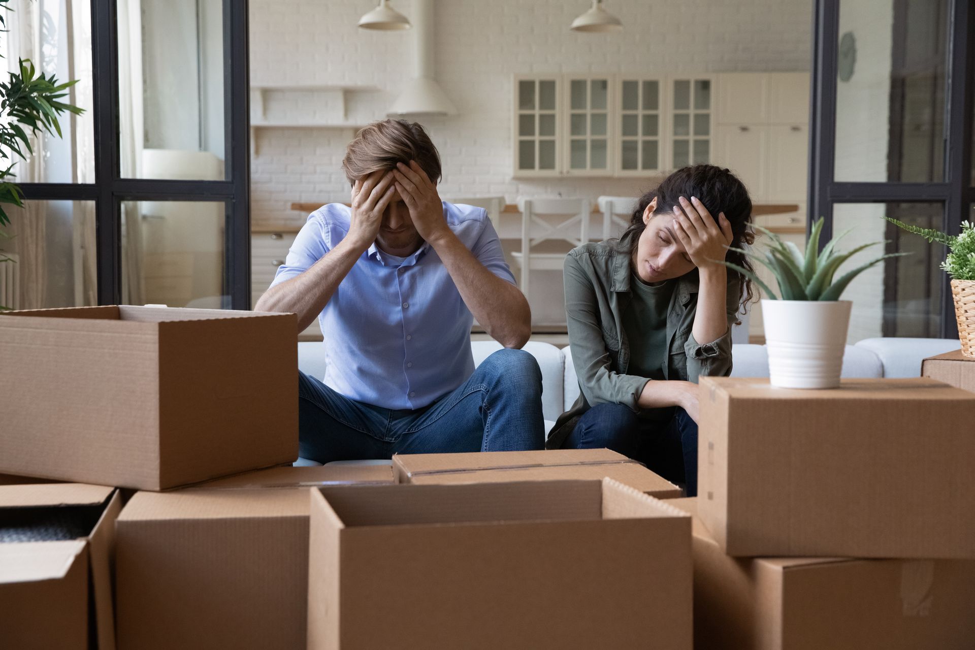 Two people sit among moving boxes, covering their faces in a bright room.