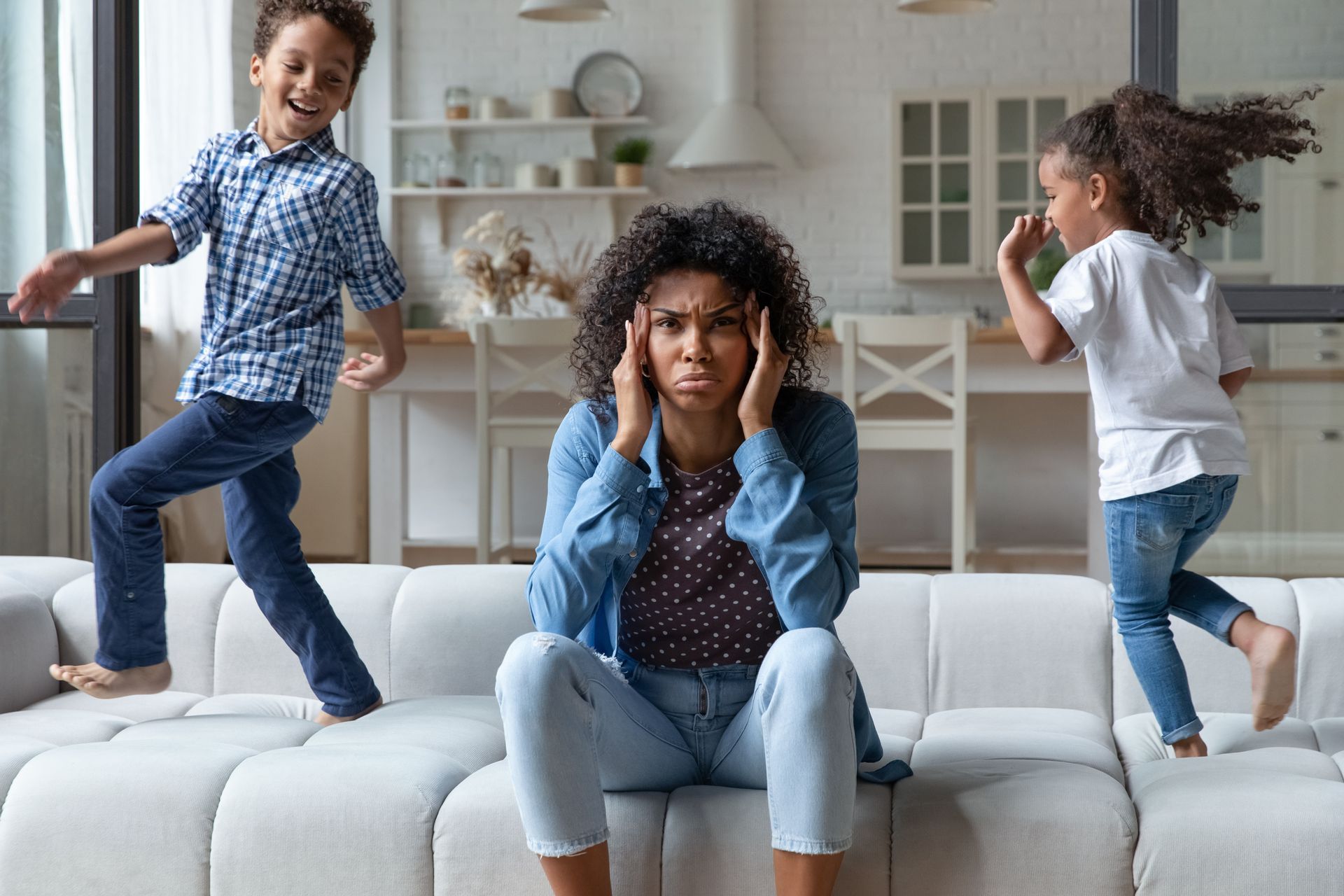Children playing on a white couch while one woman sits between them holding her head in a bright living room