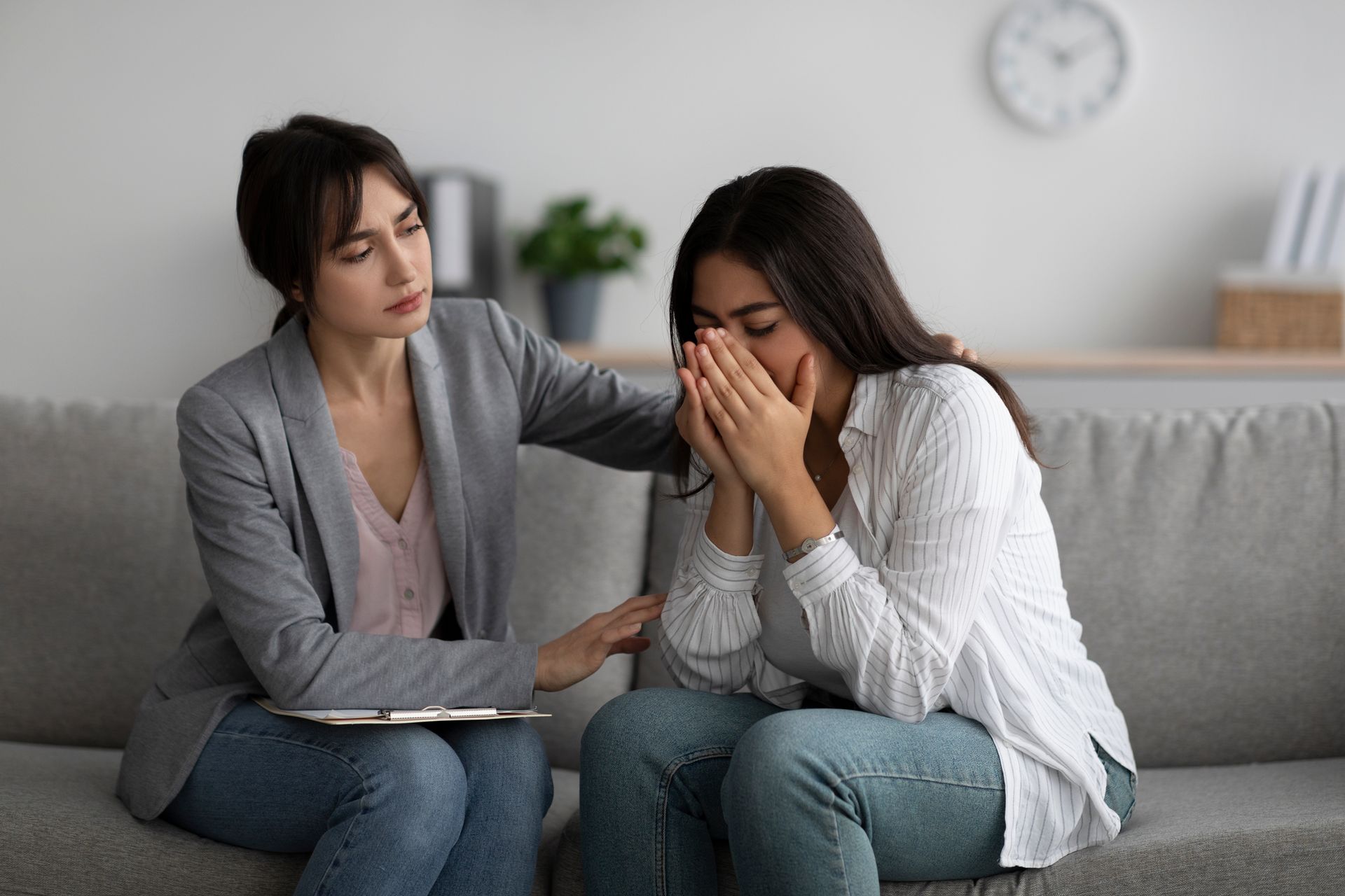 Counselor comforting a distressed woman on a gray sofa in a bright living room