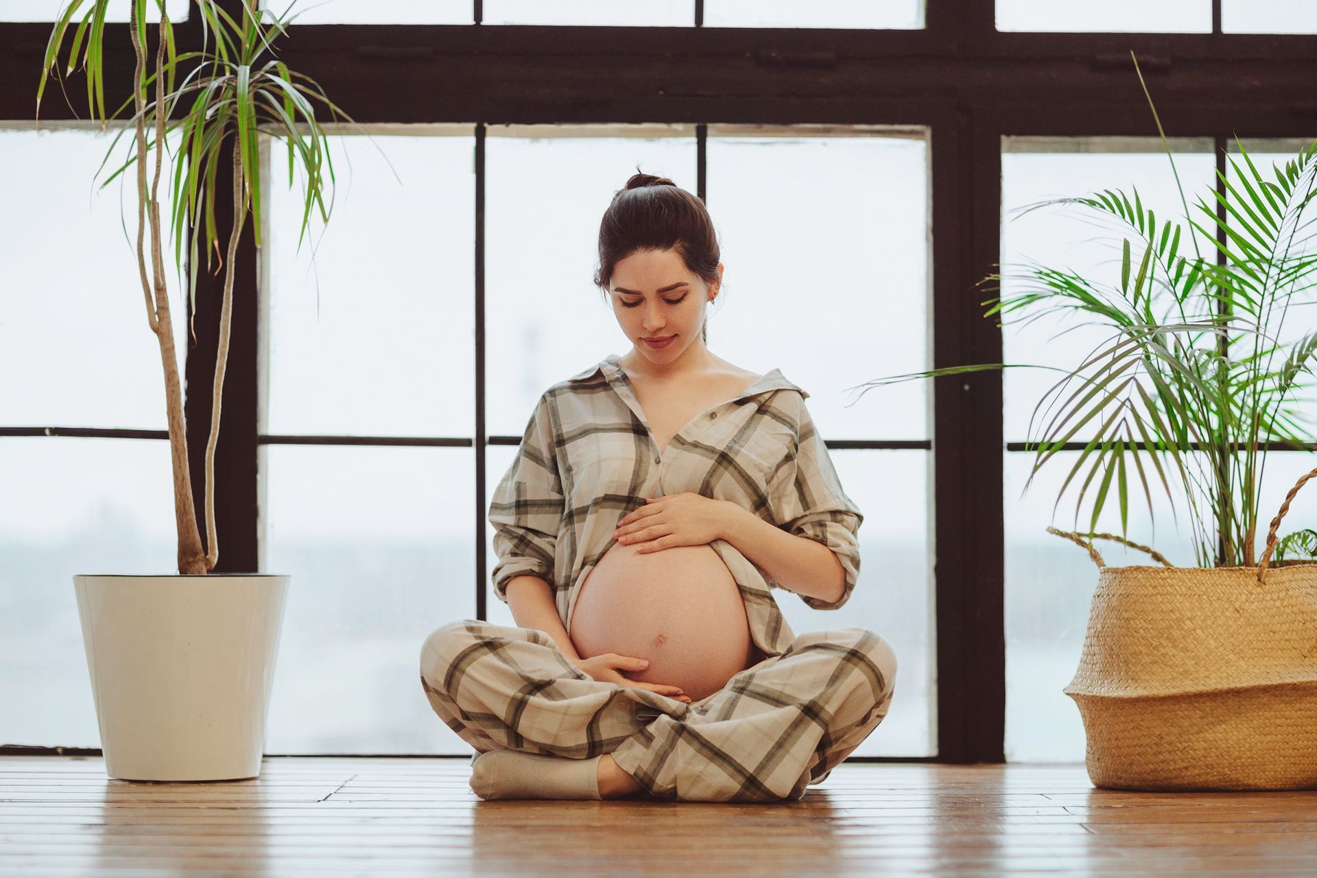 Pregnant person sitting cross-legged indoors near large windows, holding their belly, with potted plants nearby