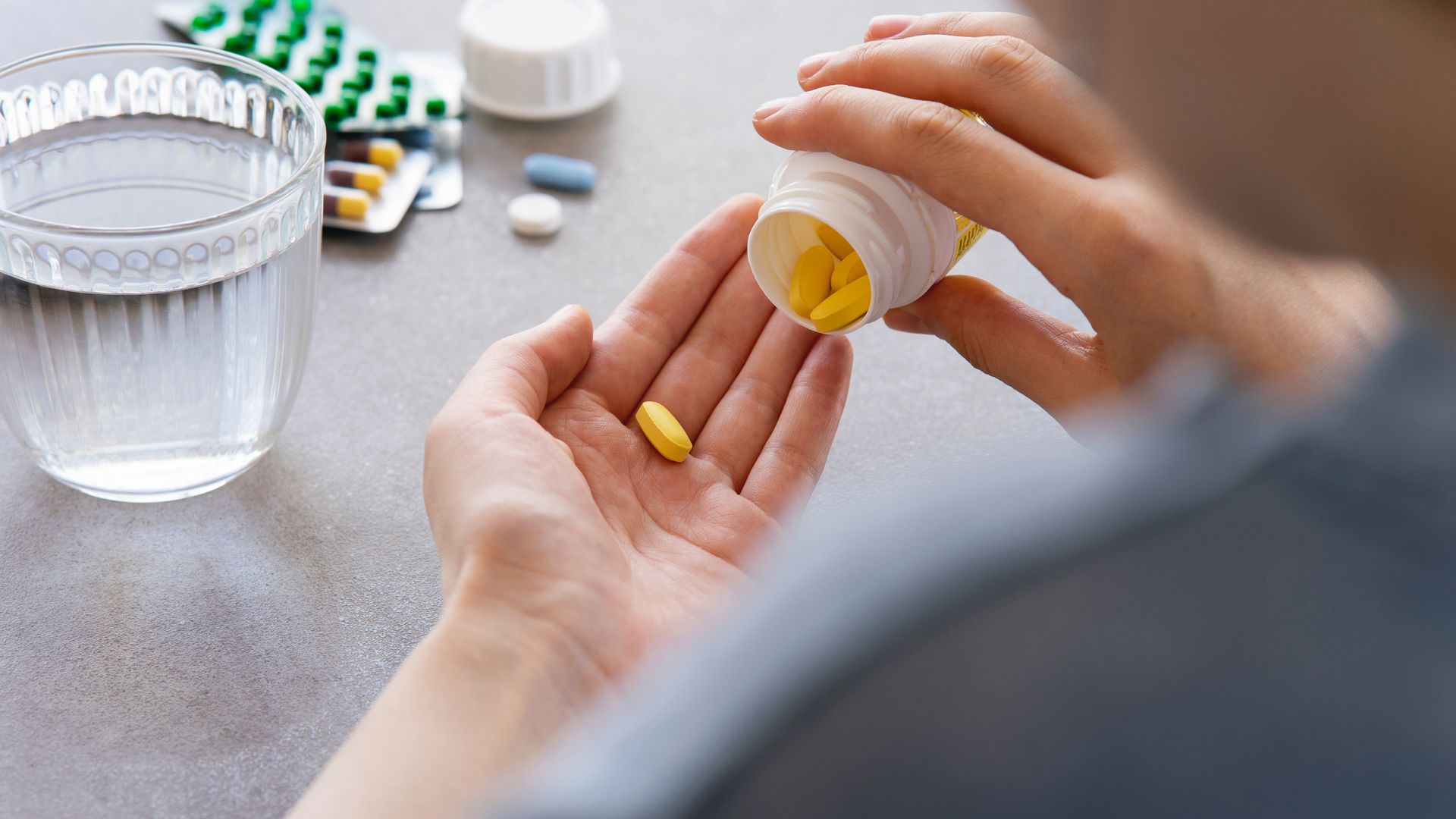 Hand pouring yellow pills from a bottle into an open palm beside a water glass and scattered tablets.