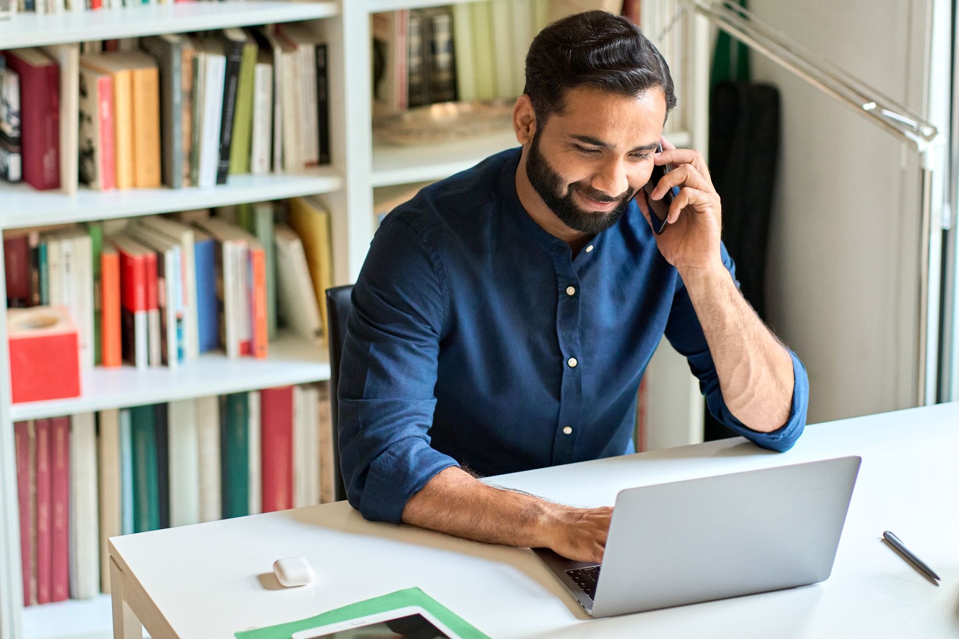 Man seated at a desk, talking on the phone while using a laptop in a bright home office