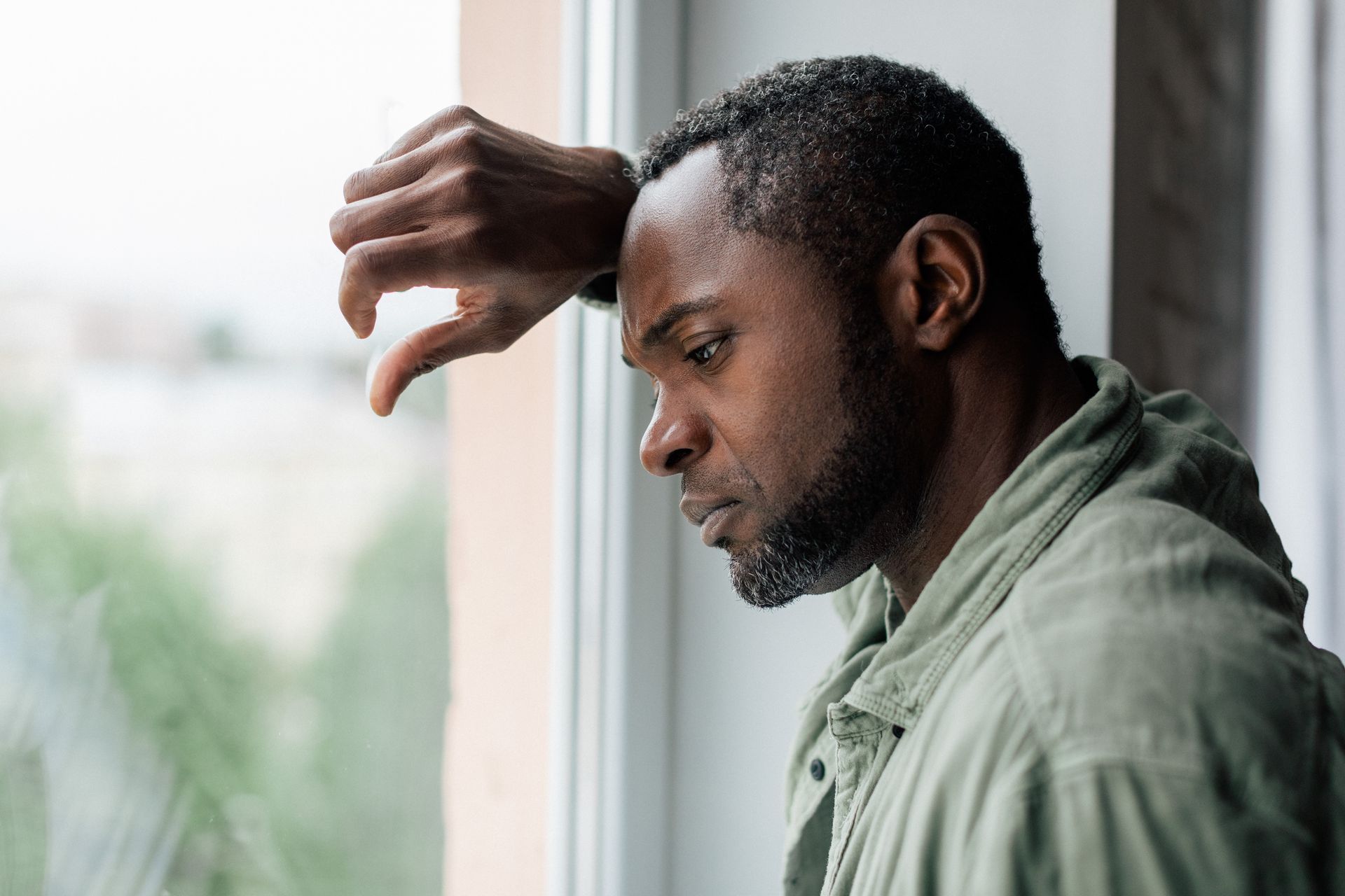 Man leaning by a window, head down with a worried expression, hand raised to his forehead.