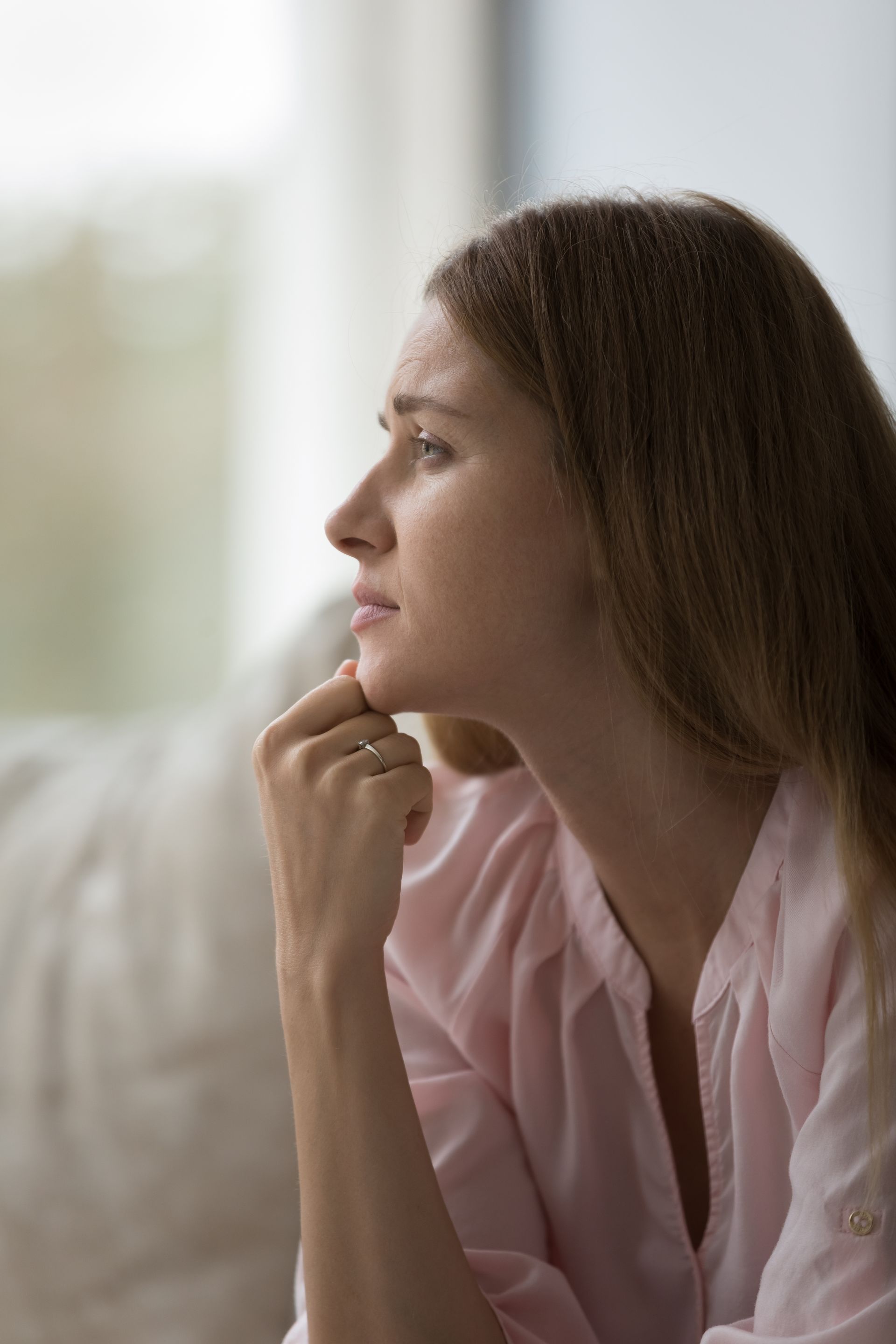 Woman in a pink blouse gazes thoughtfully out a window, resting her chin on her hand.