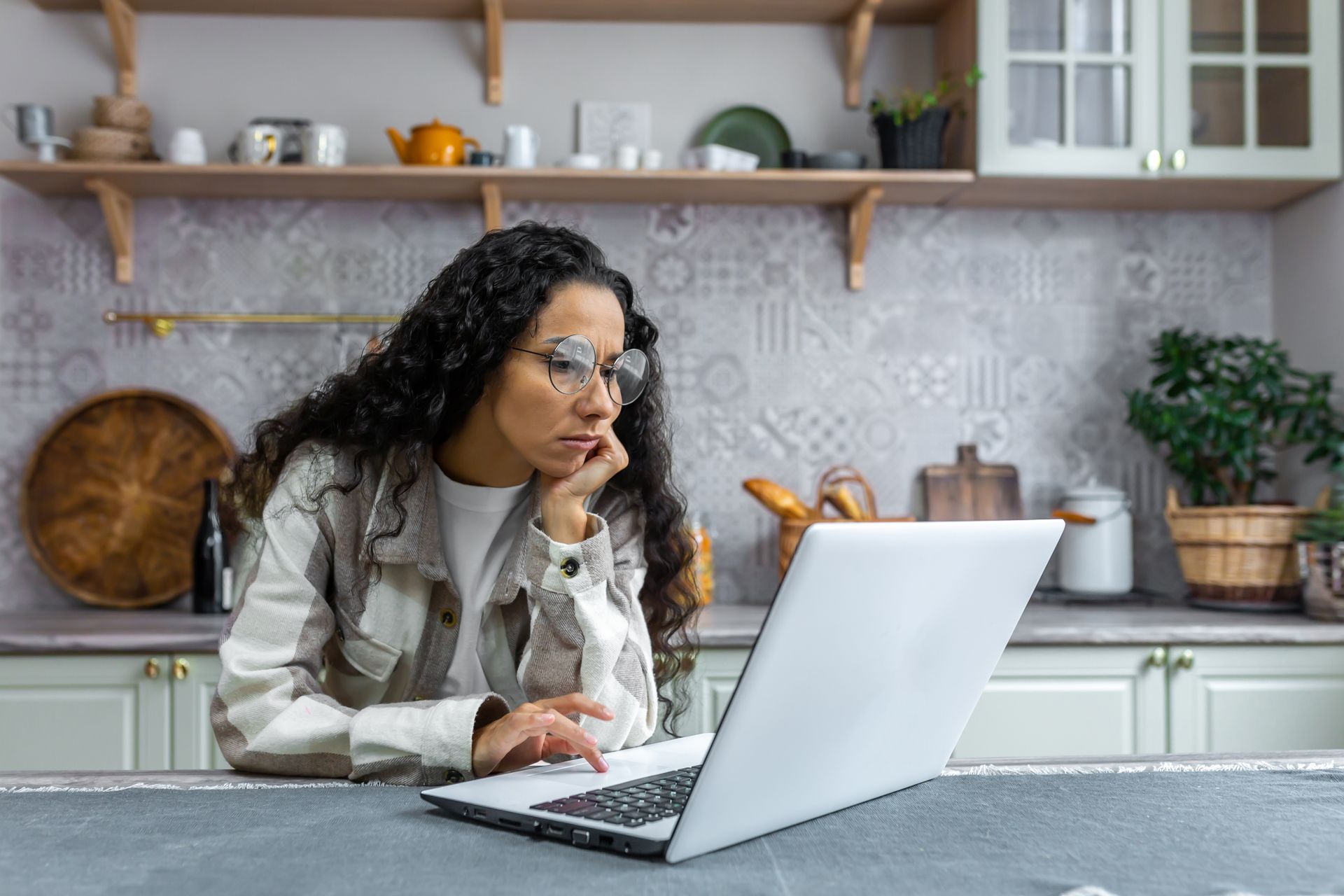 Woman working on a laptop in a bright kitchen, looking at the screen with her hand near her face.