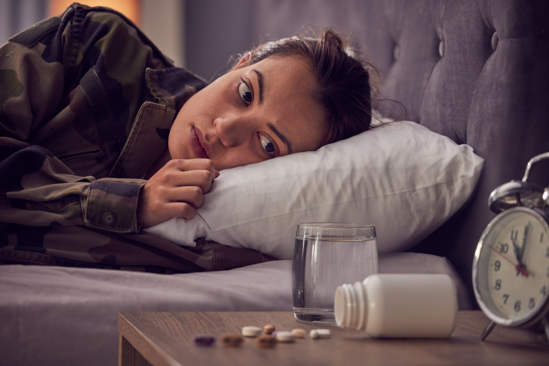 Woman lying on a pillow beside an alarm clock, medicine bottle, and pills on a bedside table