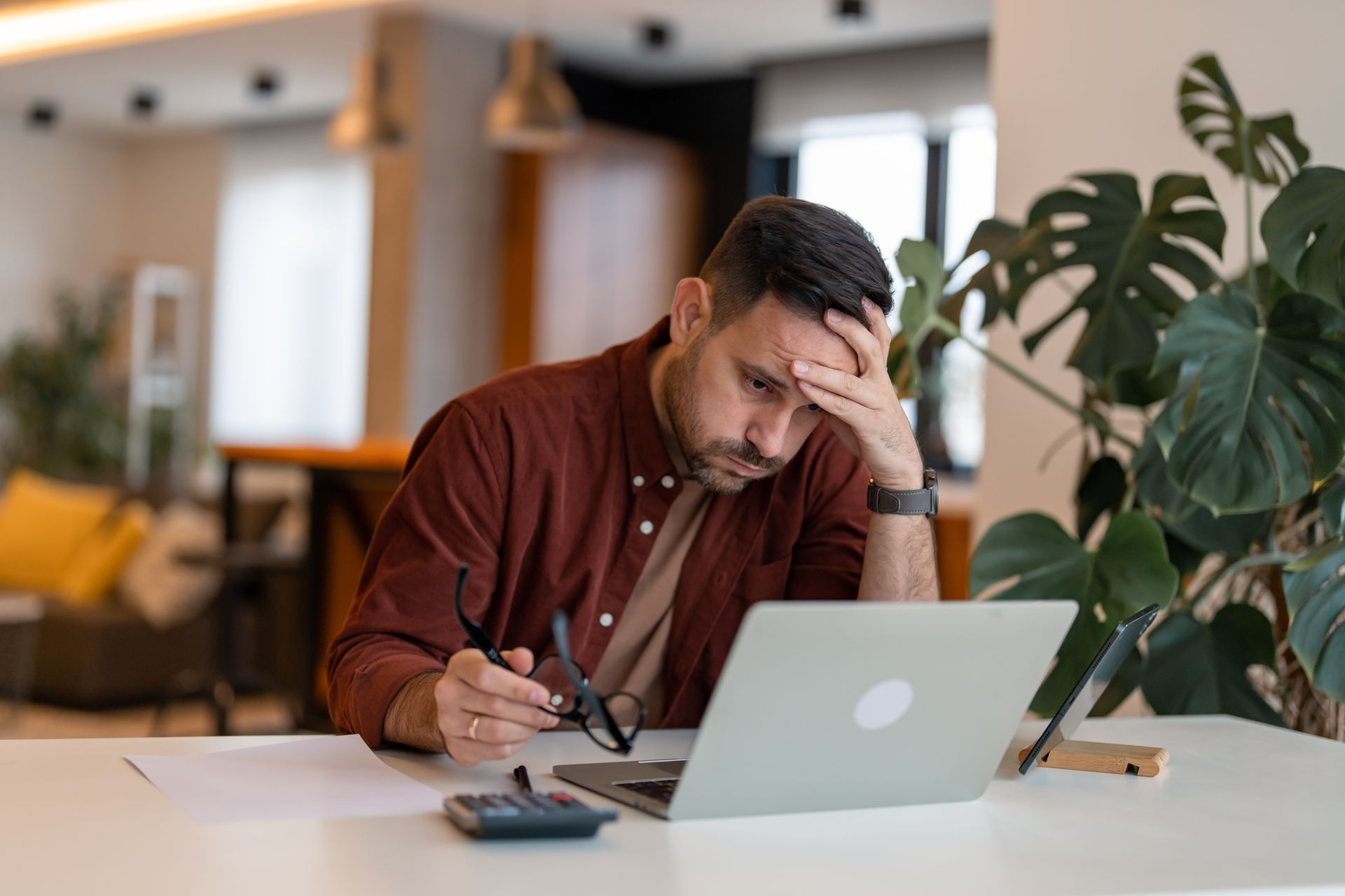 Man at a desk with a laptop, holding his forehead and looking stressed while working in a modern office