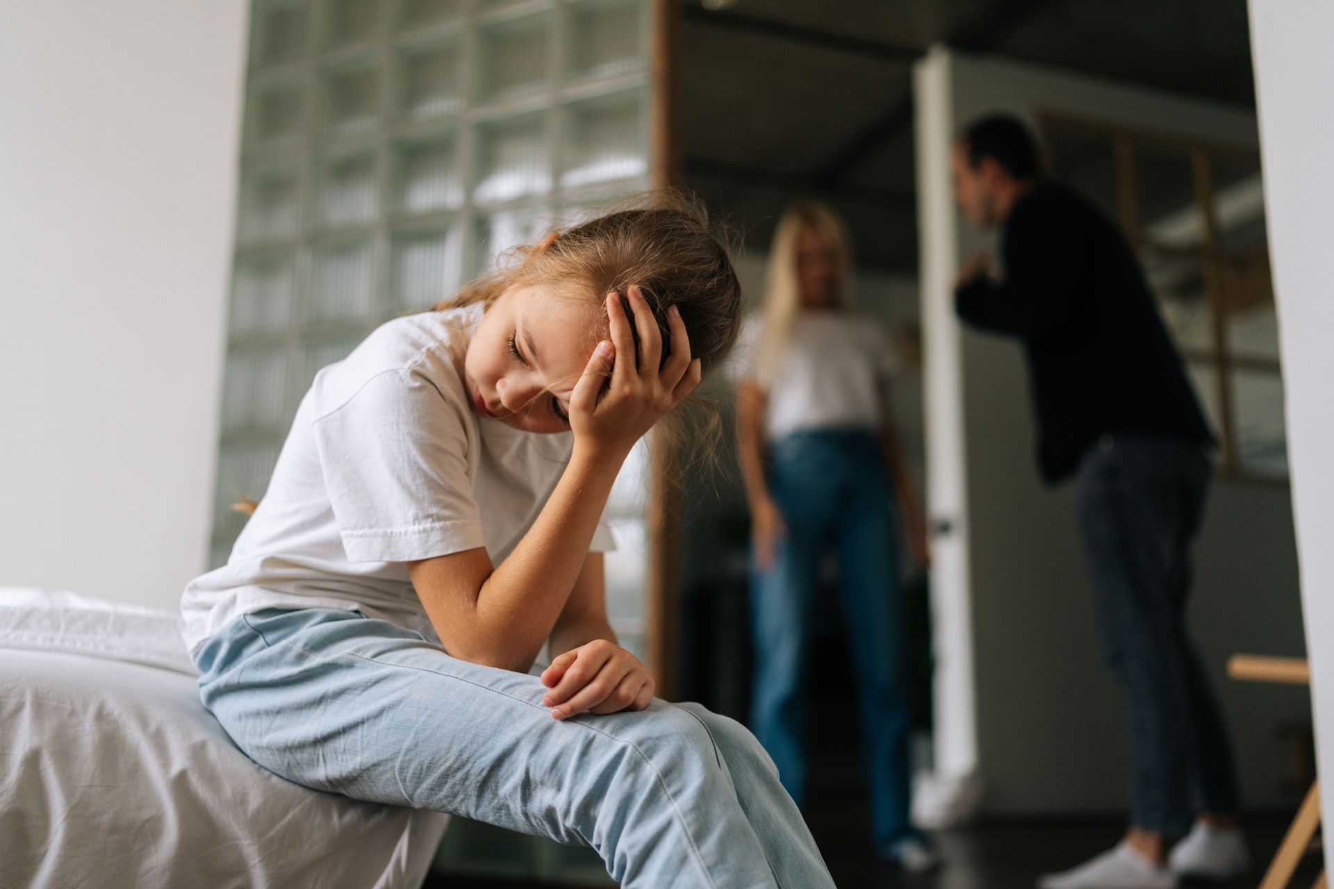 Distressed child sitting on a bed, covering her face while adults talk in the background