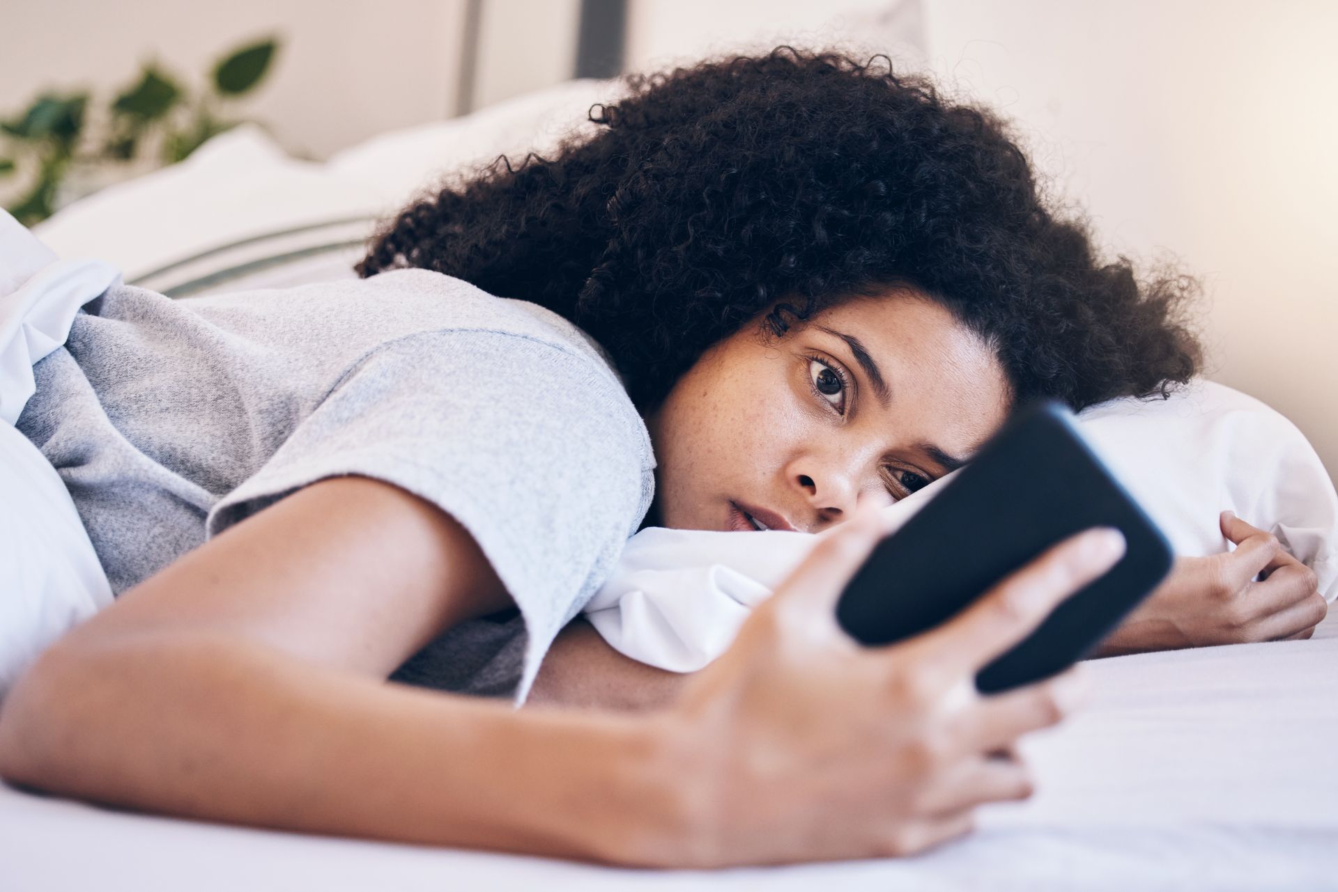 Person lying on a bed, looking at a smartphone with a thoughtful expression.