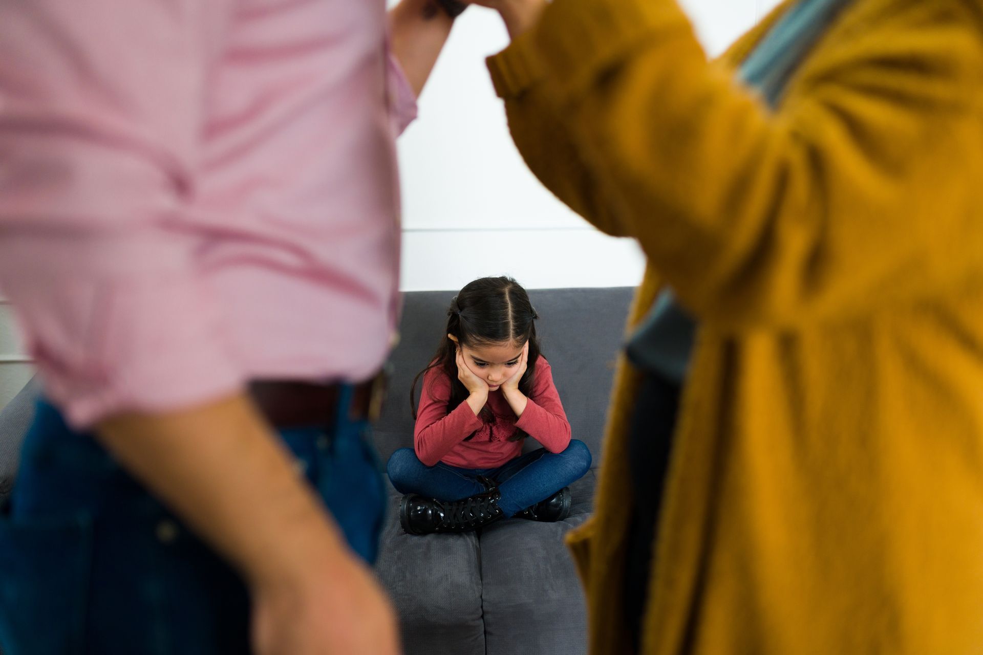 Child sitting on a couch, covering their ears while two adults stand nearby in a living room