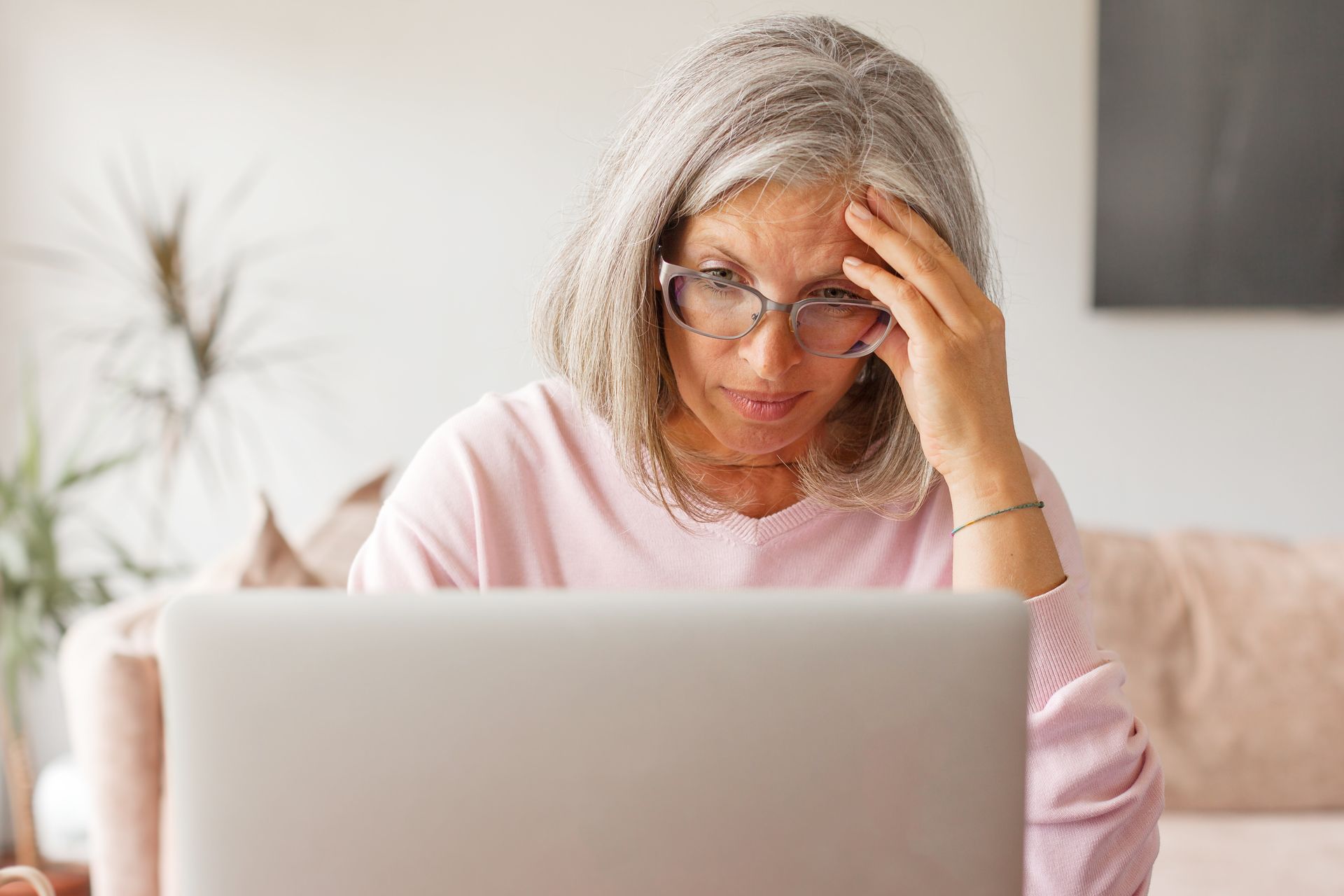 Woman in pink sweater looking stressed while using a laptop on a couch