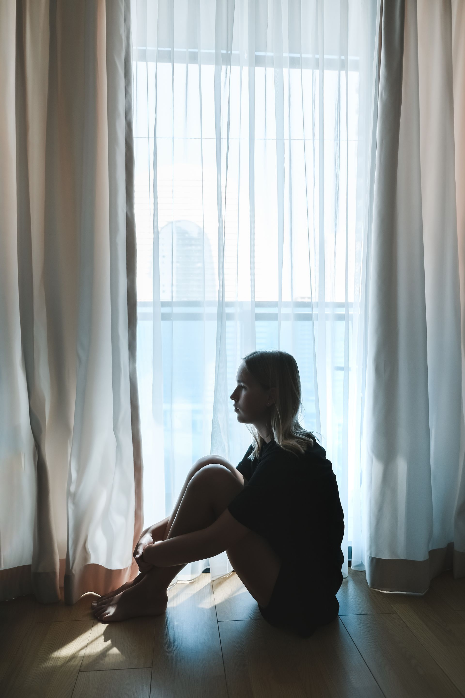 Silhouette of a seated person by sheer curtains, looking out at a bright window in a softly lit room