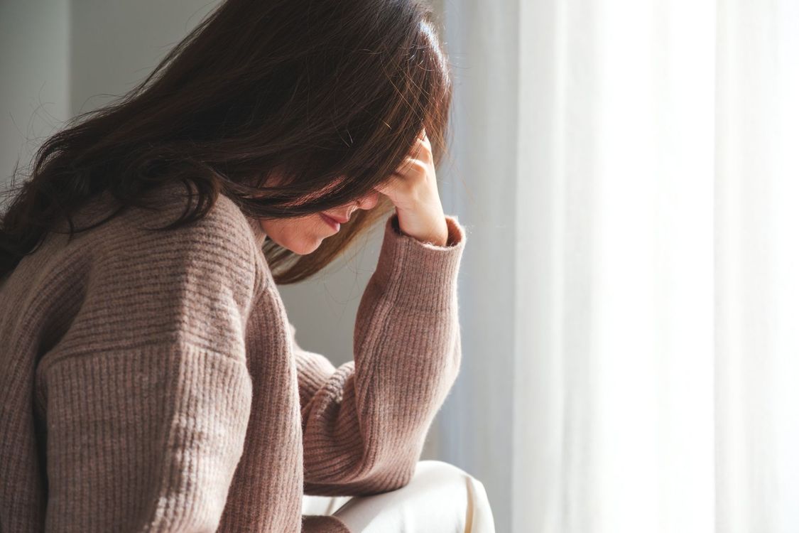 Person sitting by a window, head in hand, appearing distressed in a brown sweater