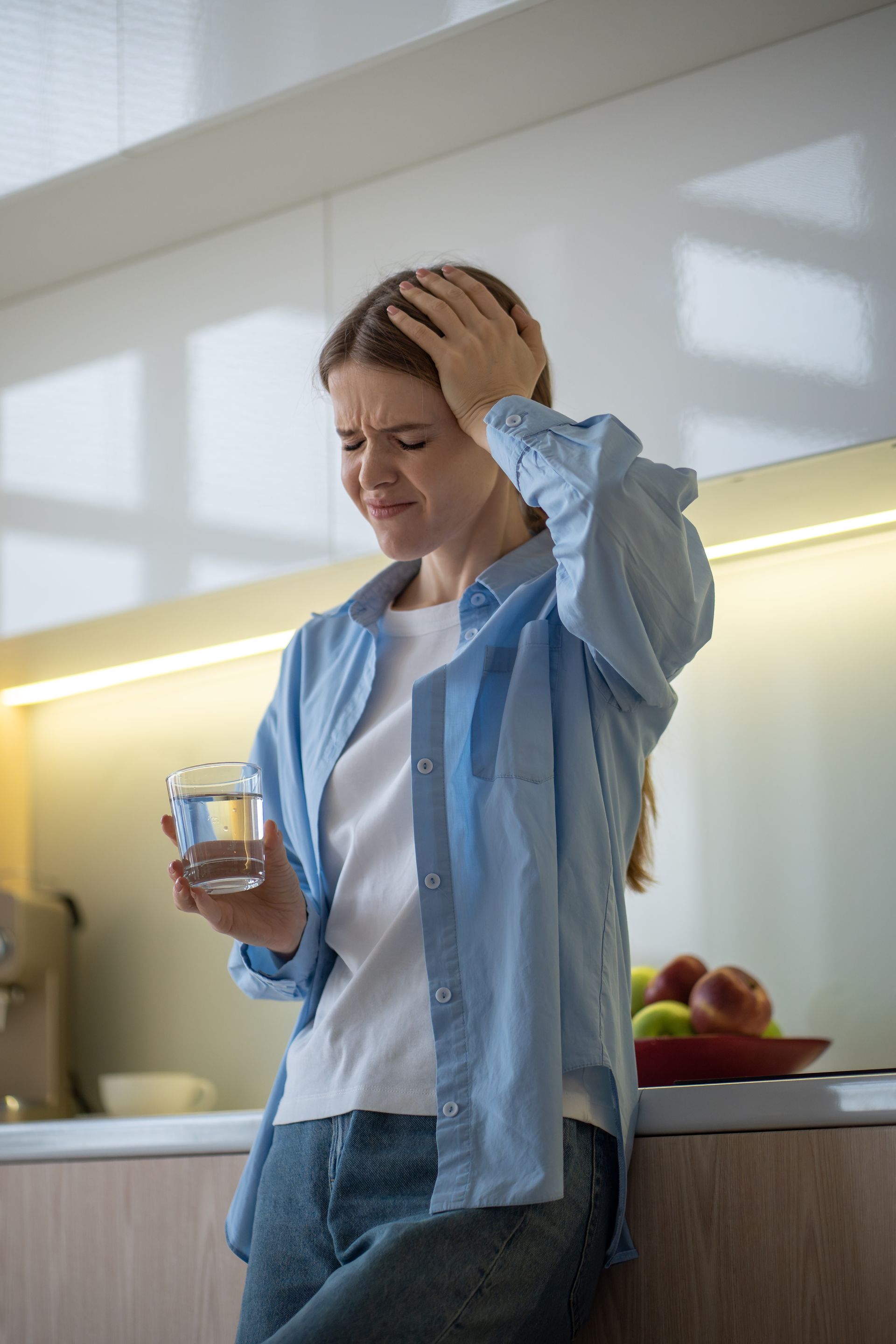 Woman in a kitchen holding a glass and rubbing her head, looking stressed.
