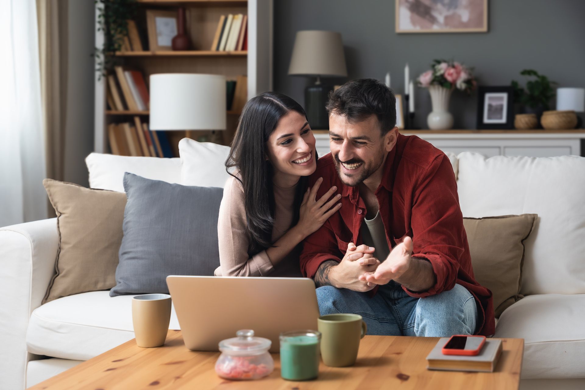 Couple smiling on a couch using a laptop, with coffee mugs and a cozy living room setting.