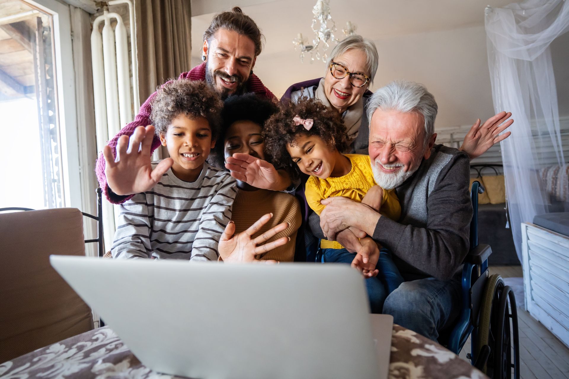 Family gathered around a laptop, smiling and hugging during a video call in a bright living room.