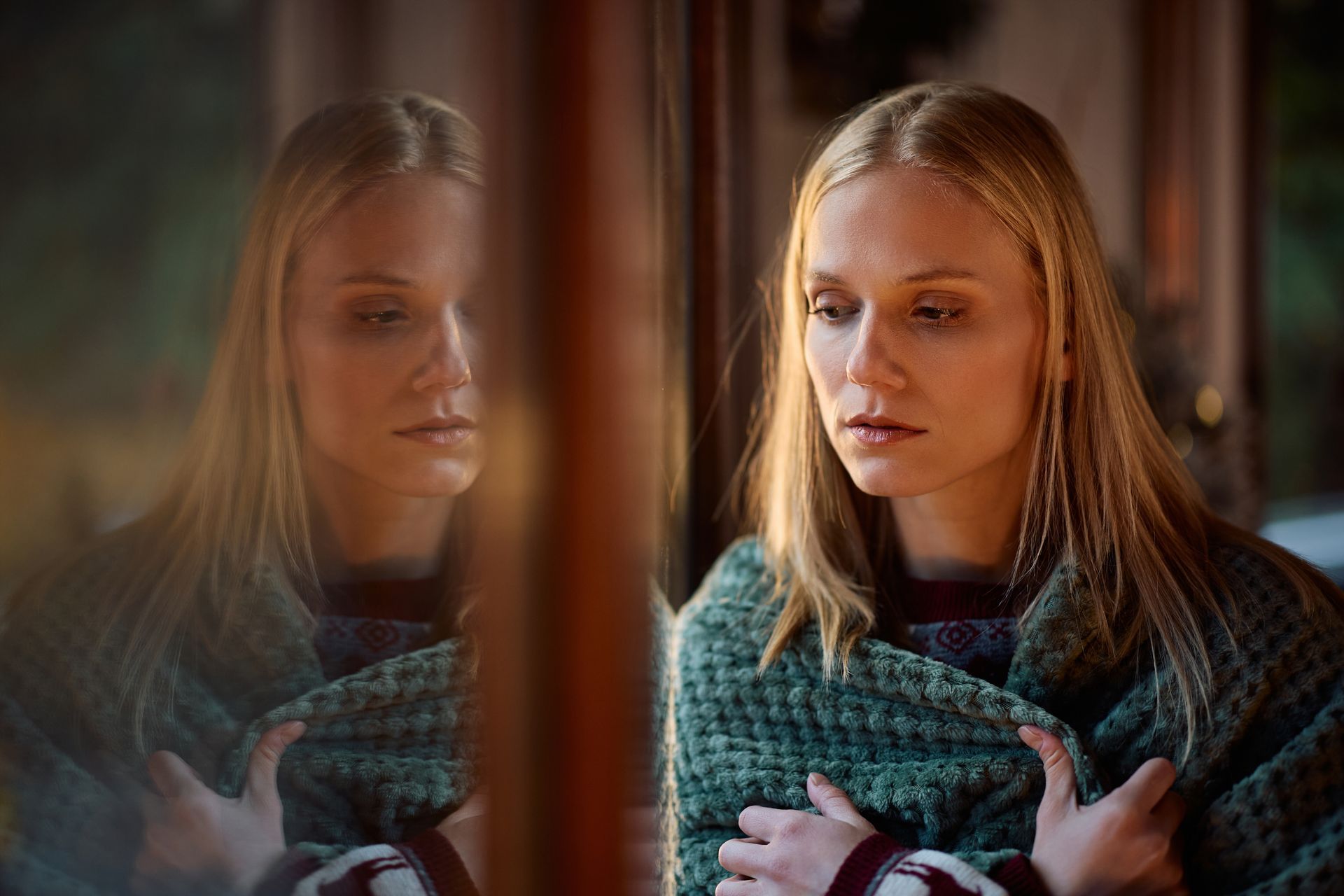 Woman in a teal sweater looking thoughtful beside a window, with her reflection visible in the glass