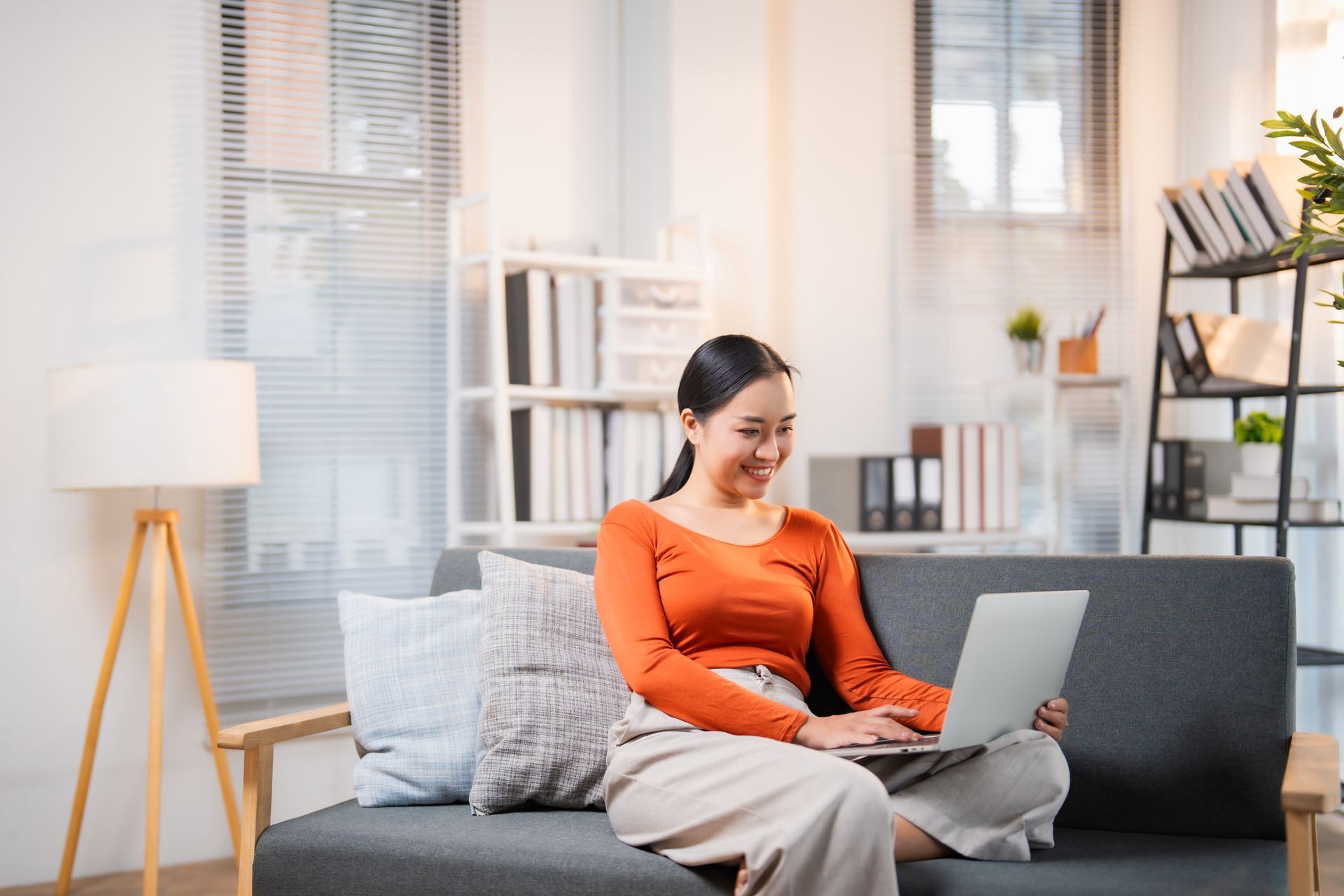 Person in an orange sweater using a laptop on a gray couch in a bright living room
