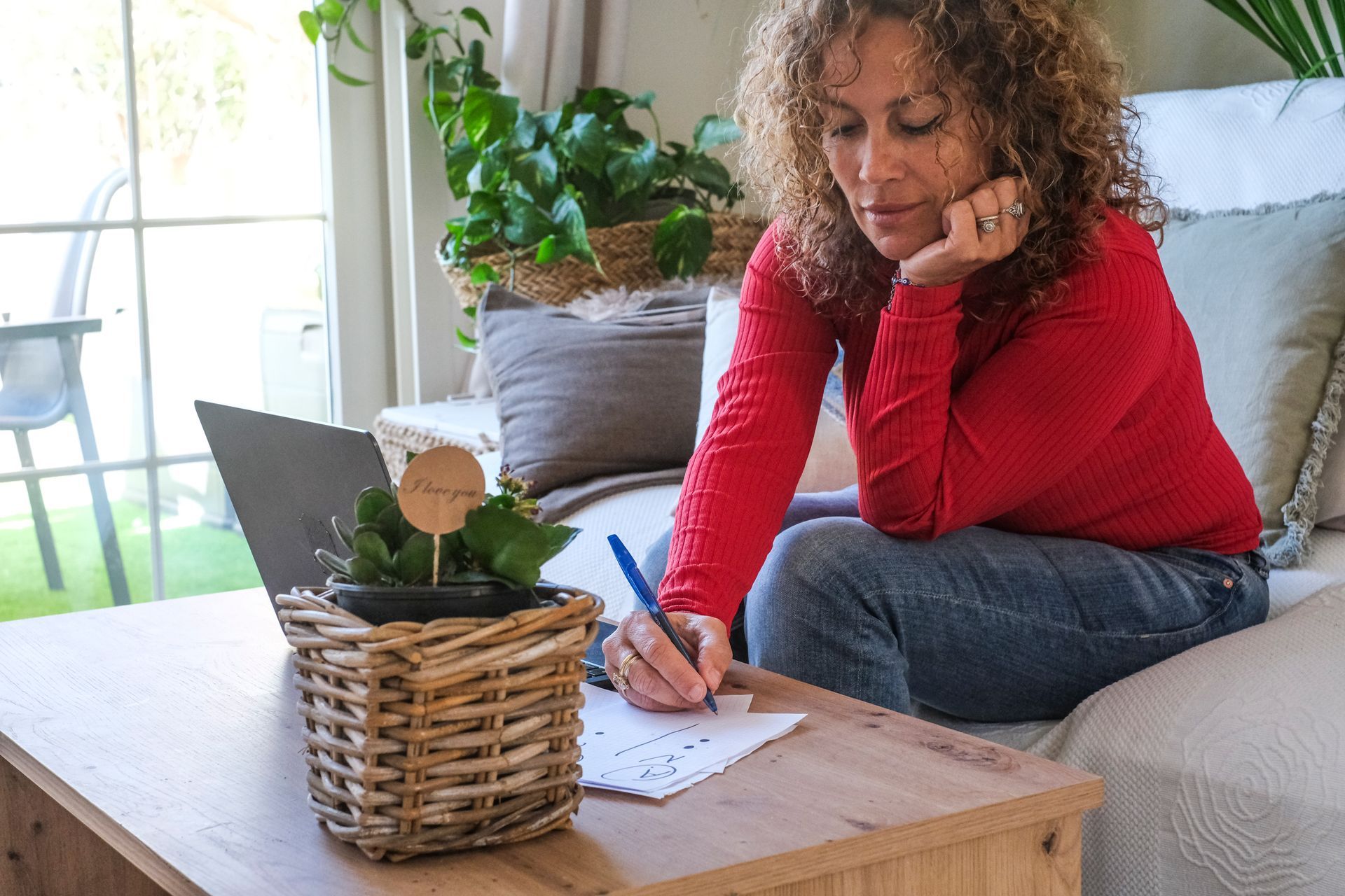 Woman in red sweater writing on paper beside a wicker planter on a living room coffee table