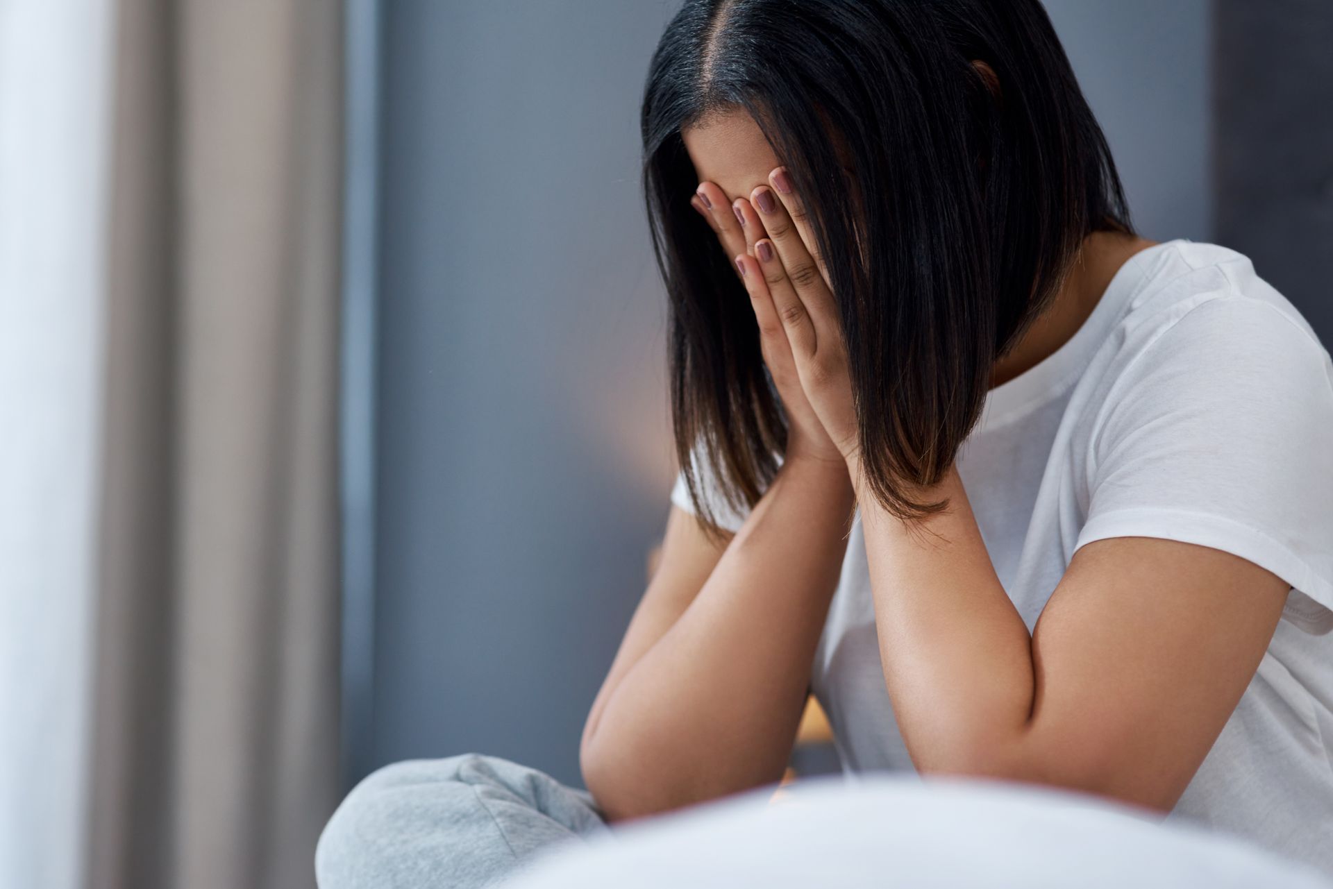 Person sitting on a bed, covering their face with hands in a dim room.