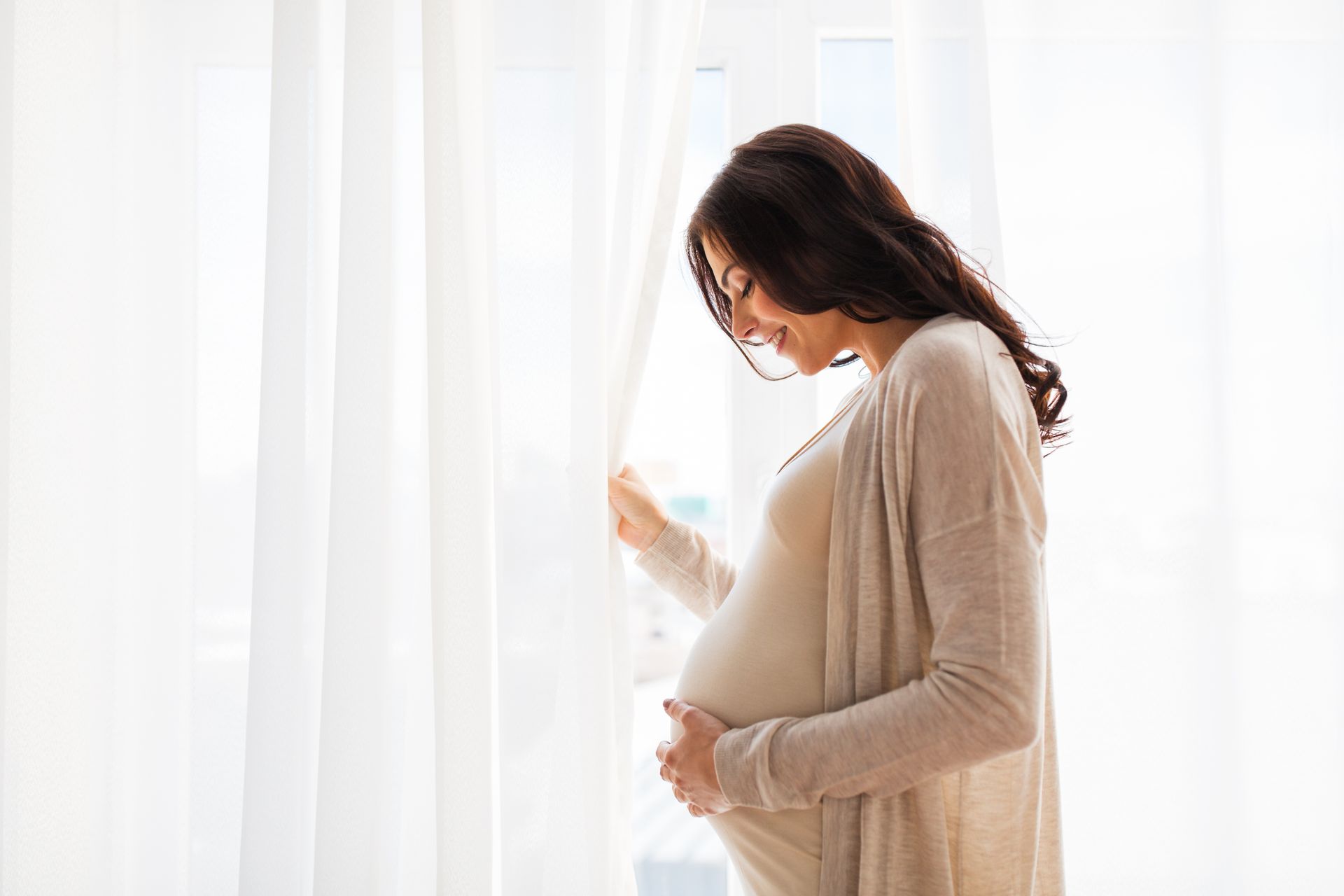 Pregnant woman standing by a bright window, gently holding her belly and curtain