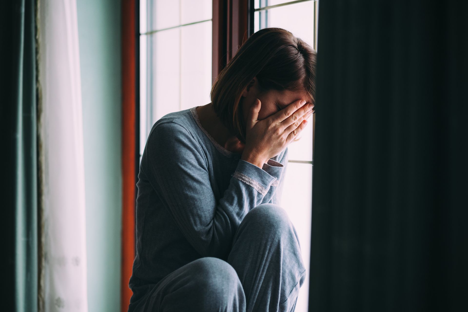 Person sitting by a window, covering their face with their hands in a dim room.