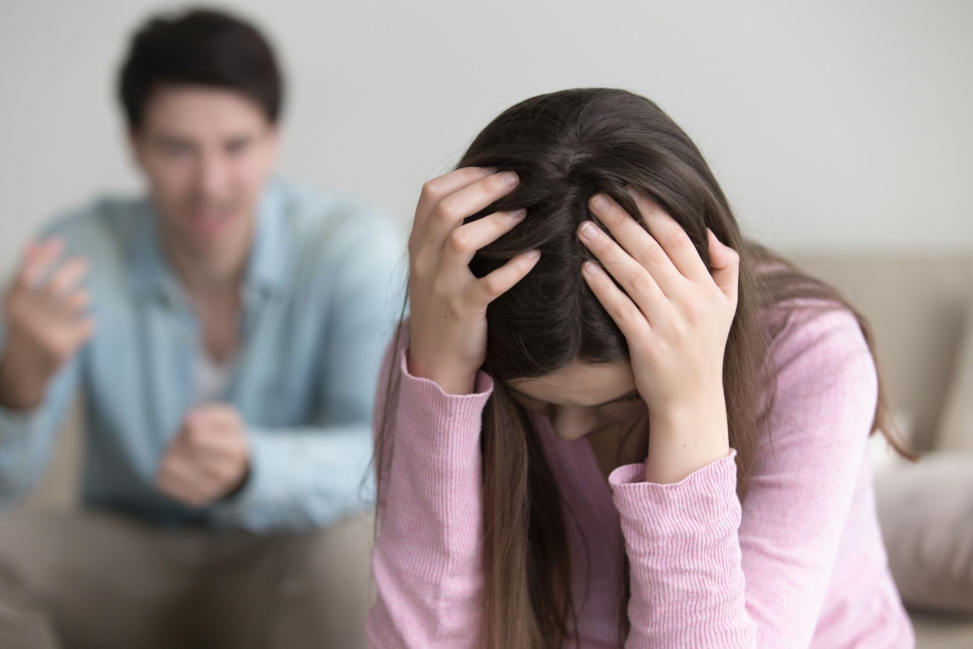 Distressed person holding head while another sits blurred in the background on a couch