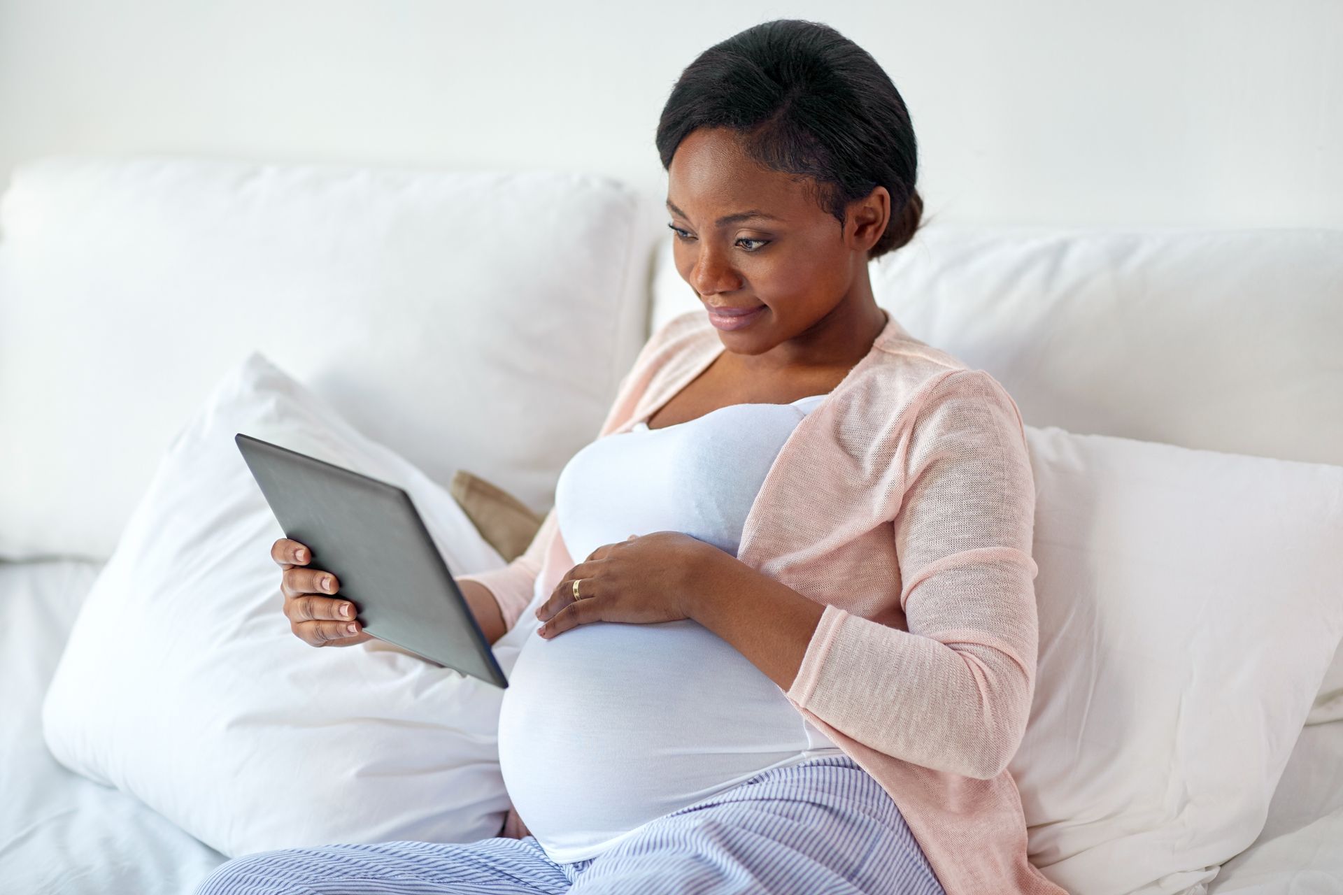 Pregnant woman sitting on a bed, looking at a tablet and resting her hand on her belly