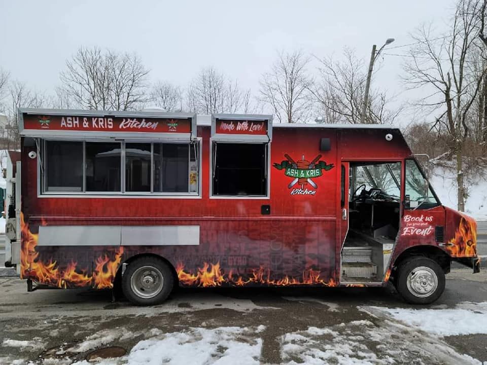Red food truck with flames, windows open, Ash & Kris Kitchen logo, parked outside.