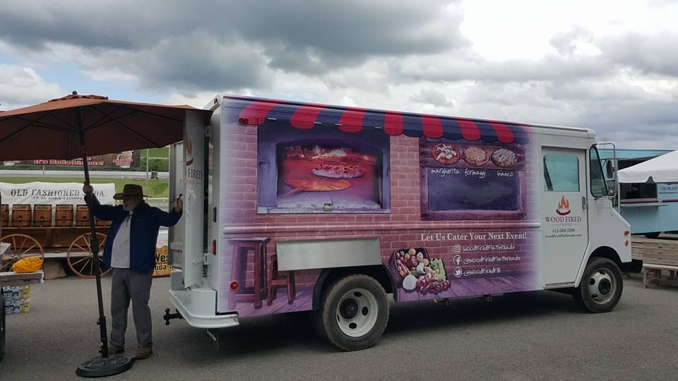 Food truck with purple brick design, person holding umbrella. White truck with awning graphic. Outdoor setting.