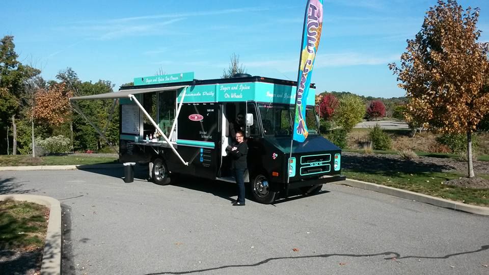 Food truck with teal and black accents, person standing at the service window, parked in a parking lot on a sunny day.