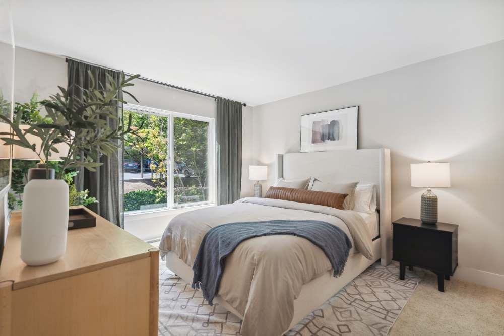 Bedroom in an apartment with a neatly made bed, two nightstands, lamps, and a large window.