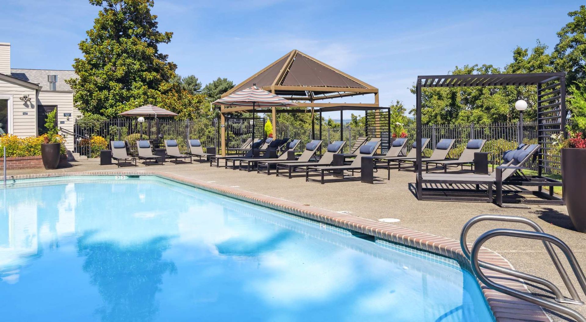 Poolside lounge area with pool, lounge chairs, pergola, and trees under a blue sky.