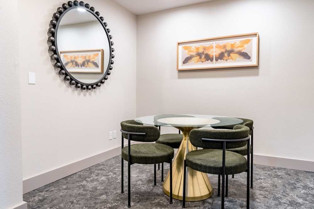 Small community lounge with a round glass table, four dark chairs, a circular bead-framed mirror, and abstract wall art.