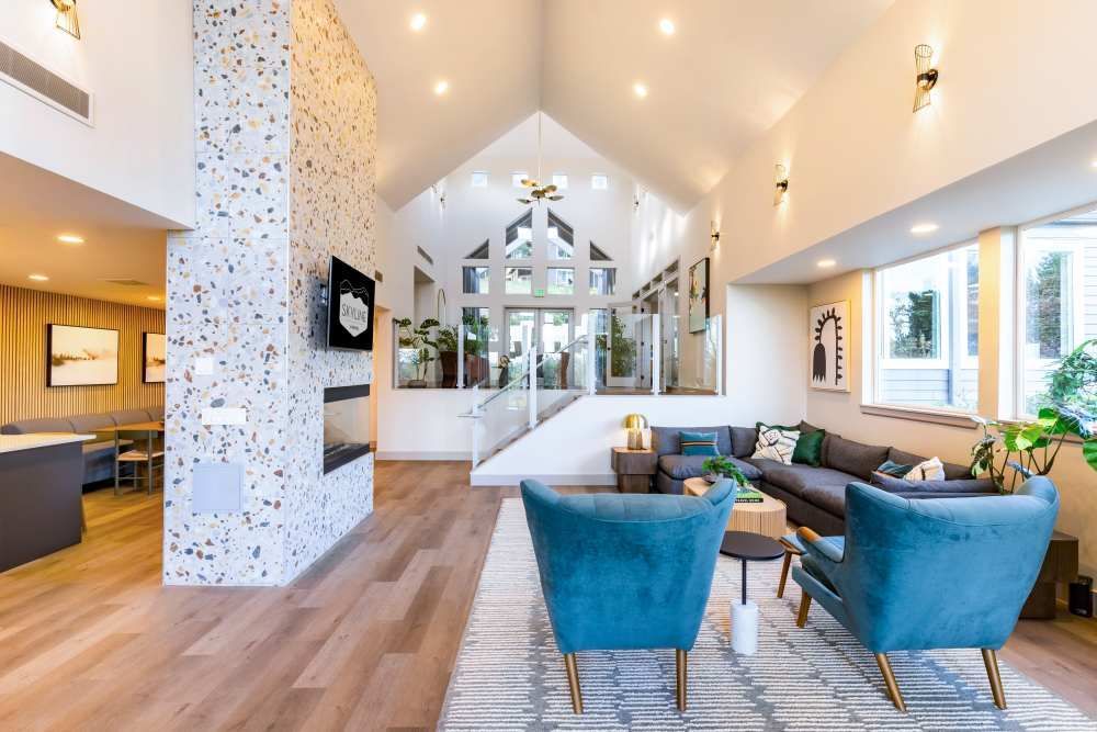 Bright interior community lobby with teal chairs, a sofa, rug, and a glass staircase.