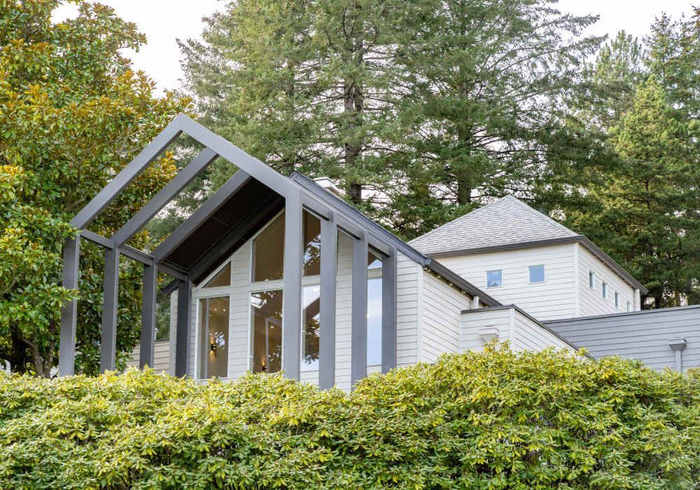 Modern white house with dark gray trim and a large glass window, surrounded by greenery.