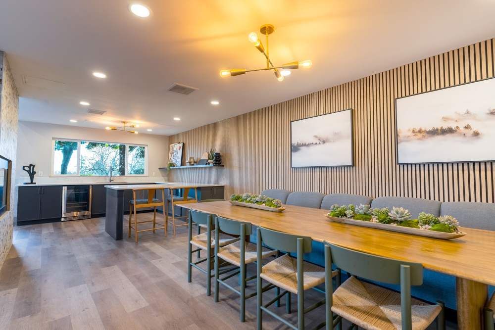 Bright communal dining and lounge area with a long wooden table, chairs, and a kitchen bar.