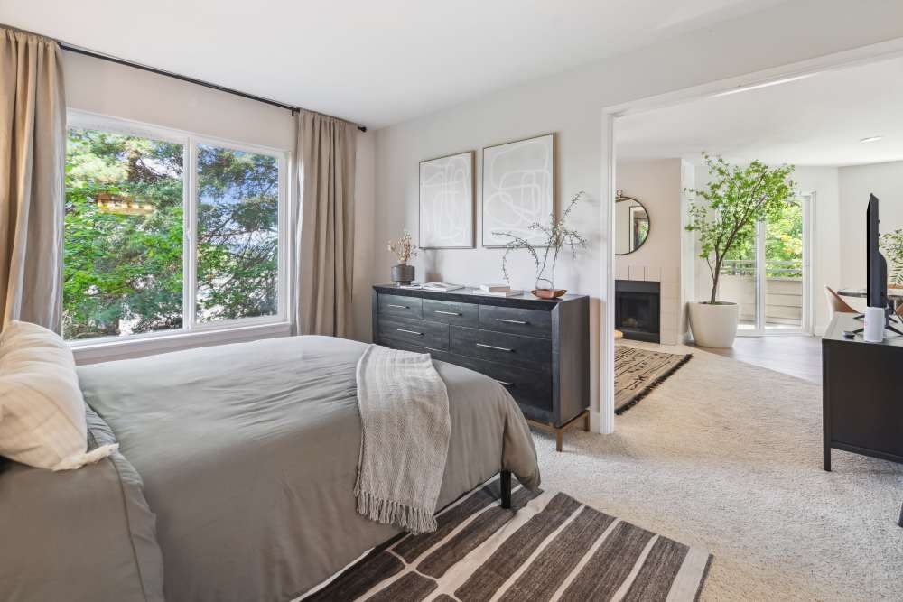Bright bedroom in a modern apartment with a large window, dresser, and open doorway to the living area.