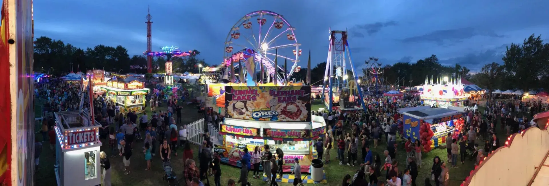 A large crowd of people are gathered at a carnival with a ferris wheel in the background.