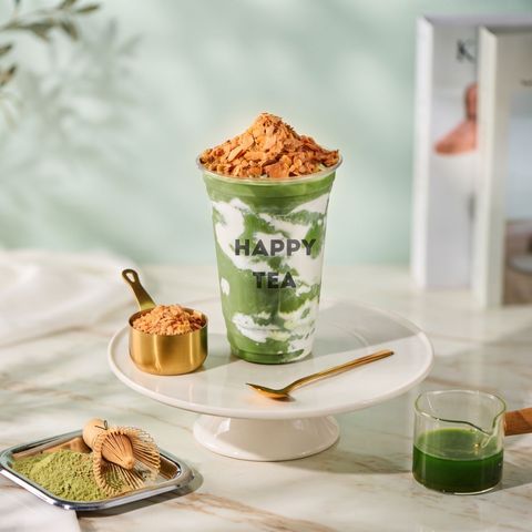 Green matcha drink topped with crumbs on a white pedestal, with matching tea items beside it