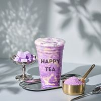 Purple “HAPPY TEA” drink with lavender foam, beside a bowl of purple cubes and a brass scoop on a gray table.