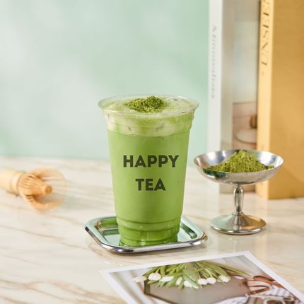Green matcha latte in a clear cup labeled “HAPPY TEA” beside a small dish of matcha powder