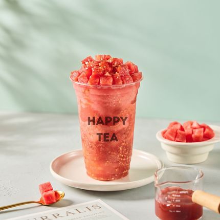 Pink drink topped with fruit cubes, labeled “HAPPY TEA,” on a saucer with a small bowl and glass nearby